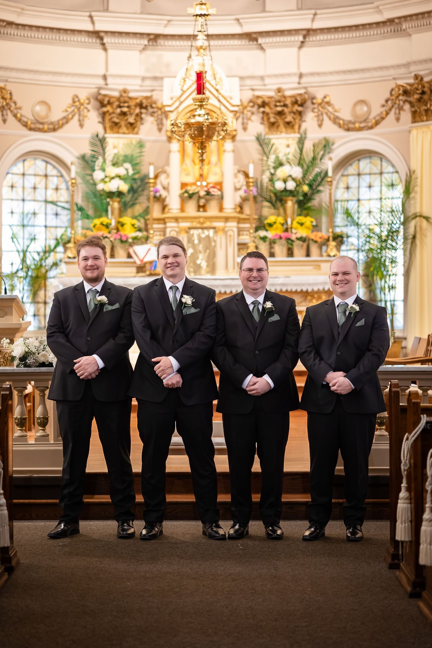 Four groomsmen in black suits with boutonnieres standing together in front of ornate church altar.