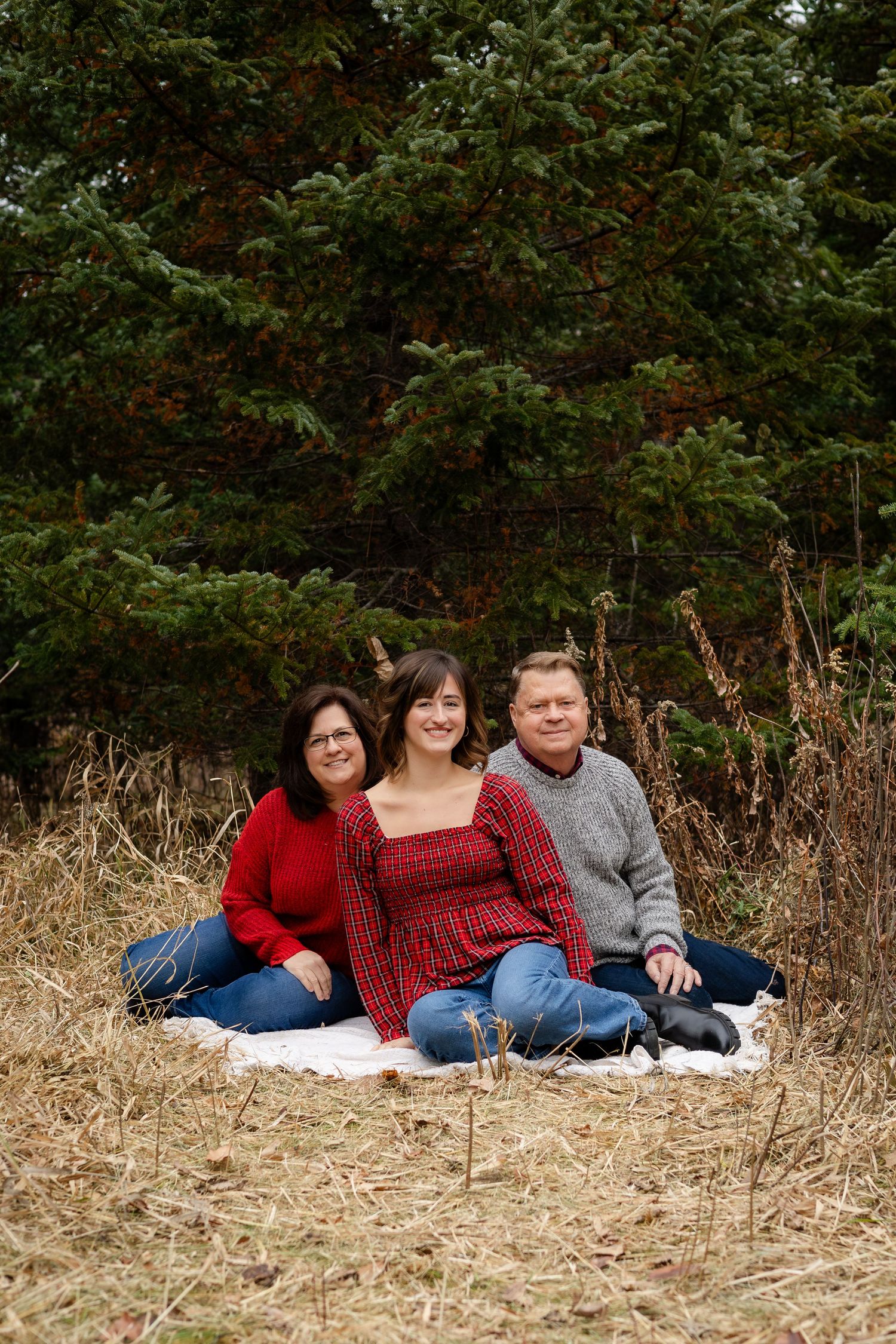 Three people in cozy winter outfits lounging on a blanket outdoors with natural scenery behind them.