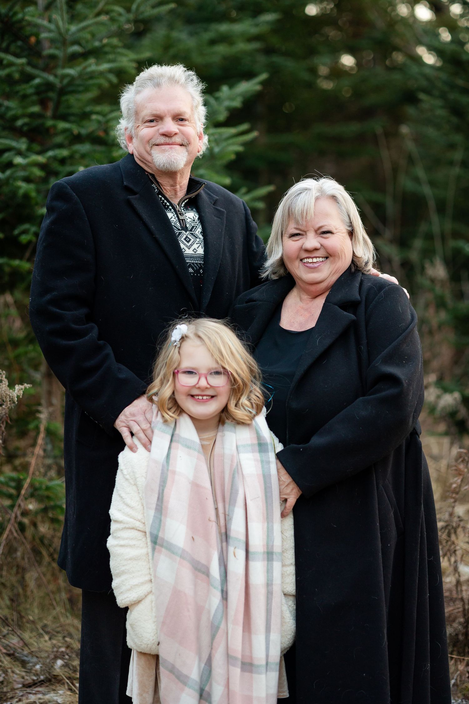 A family of three poses outdoors in winter coats against an evergreen backdrop.