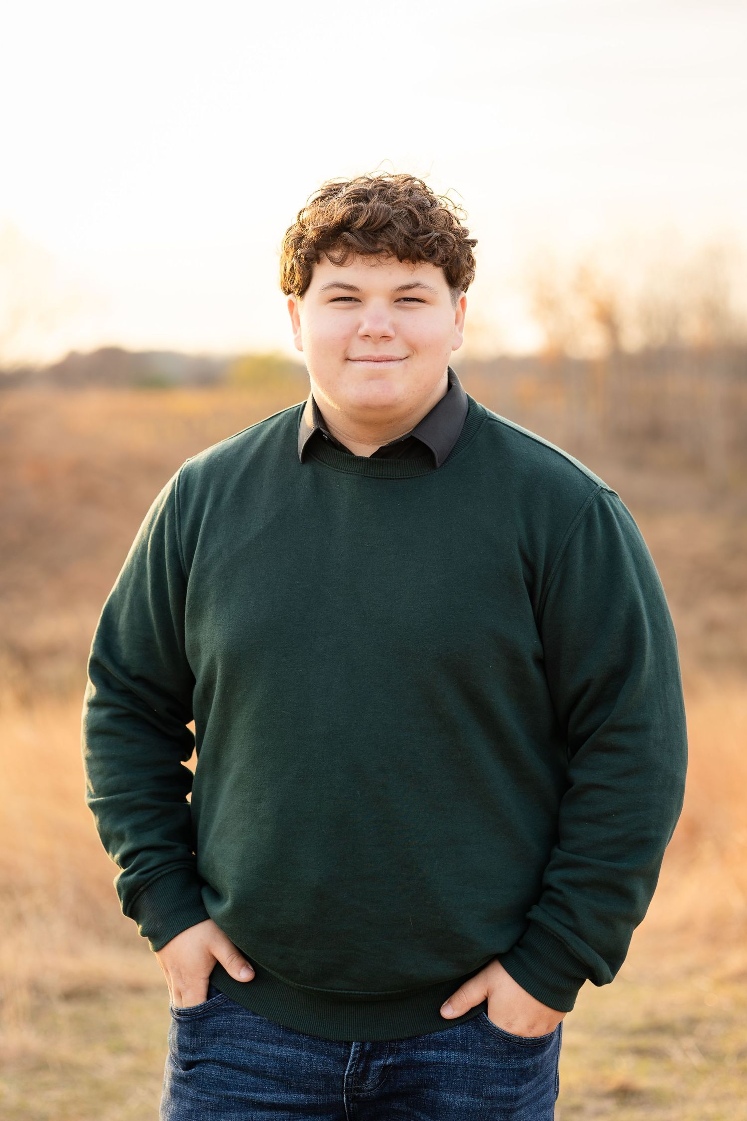 Person wearing a dark green sweater poses outdoors in a natural setting during golden hour.