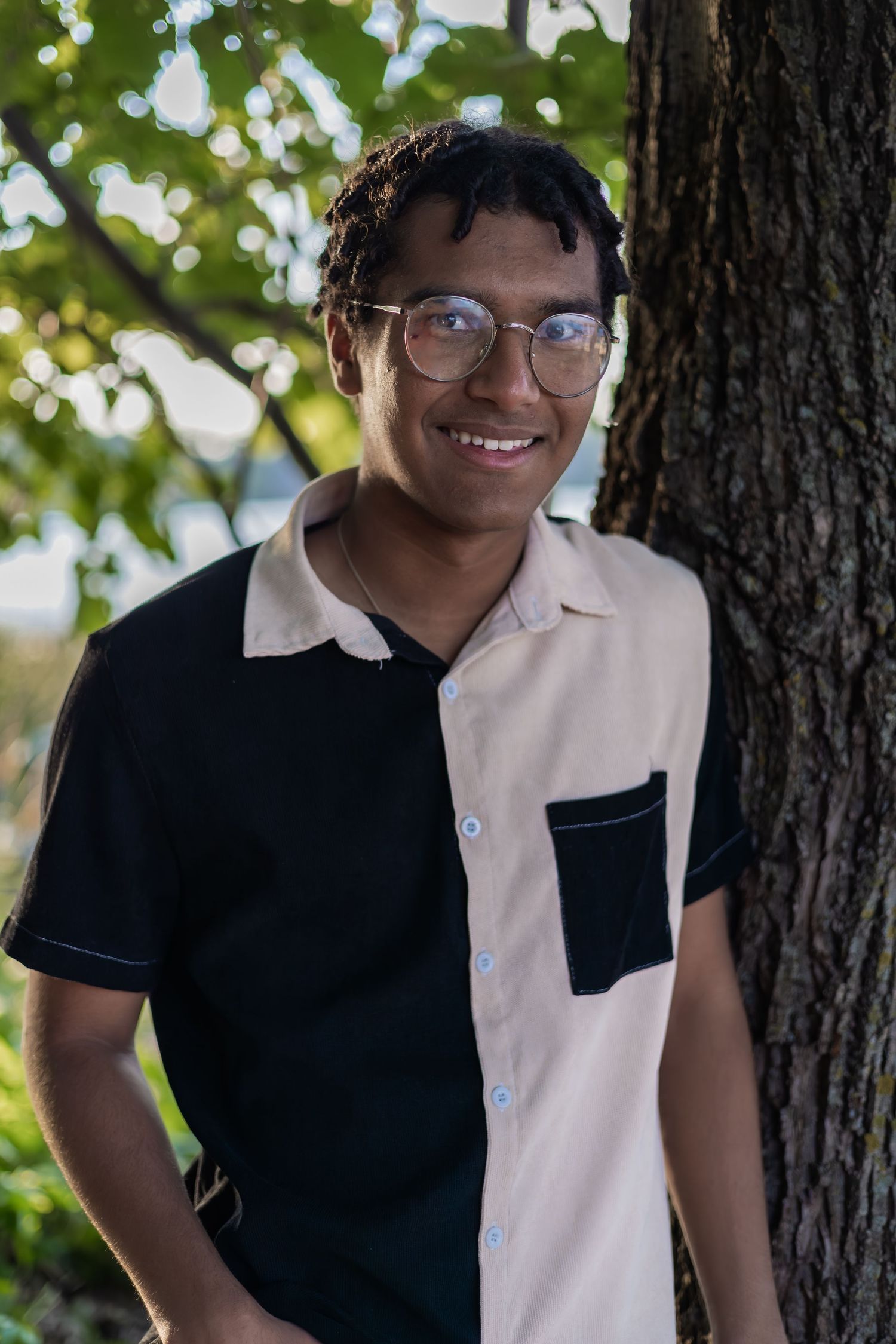 Person in two-tone shirt leaning against tree trunk with natural bokeh background.