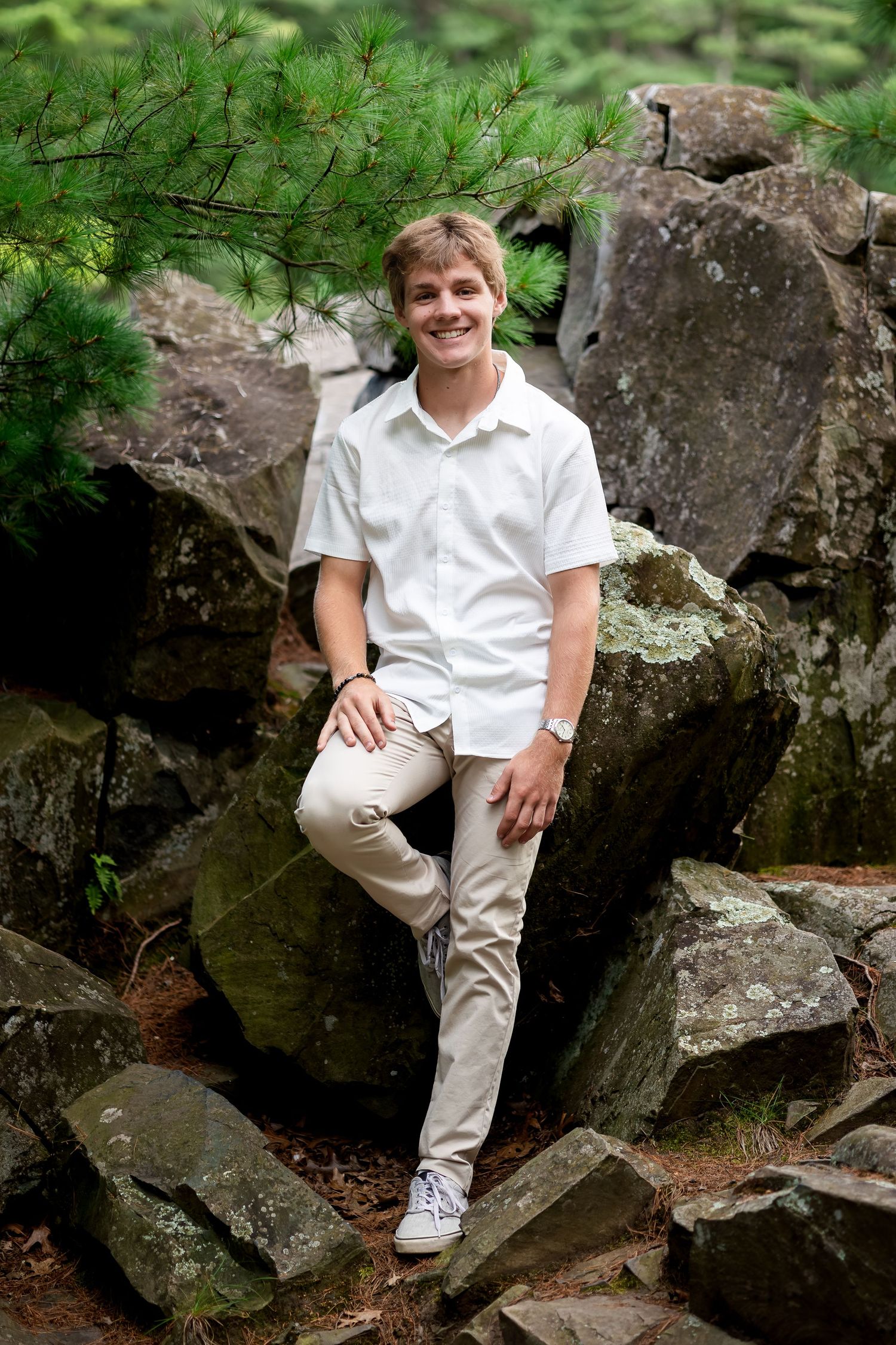 Person in all-white casual attire poses against rocky natural backdrop with greenery.
