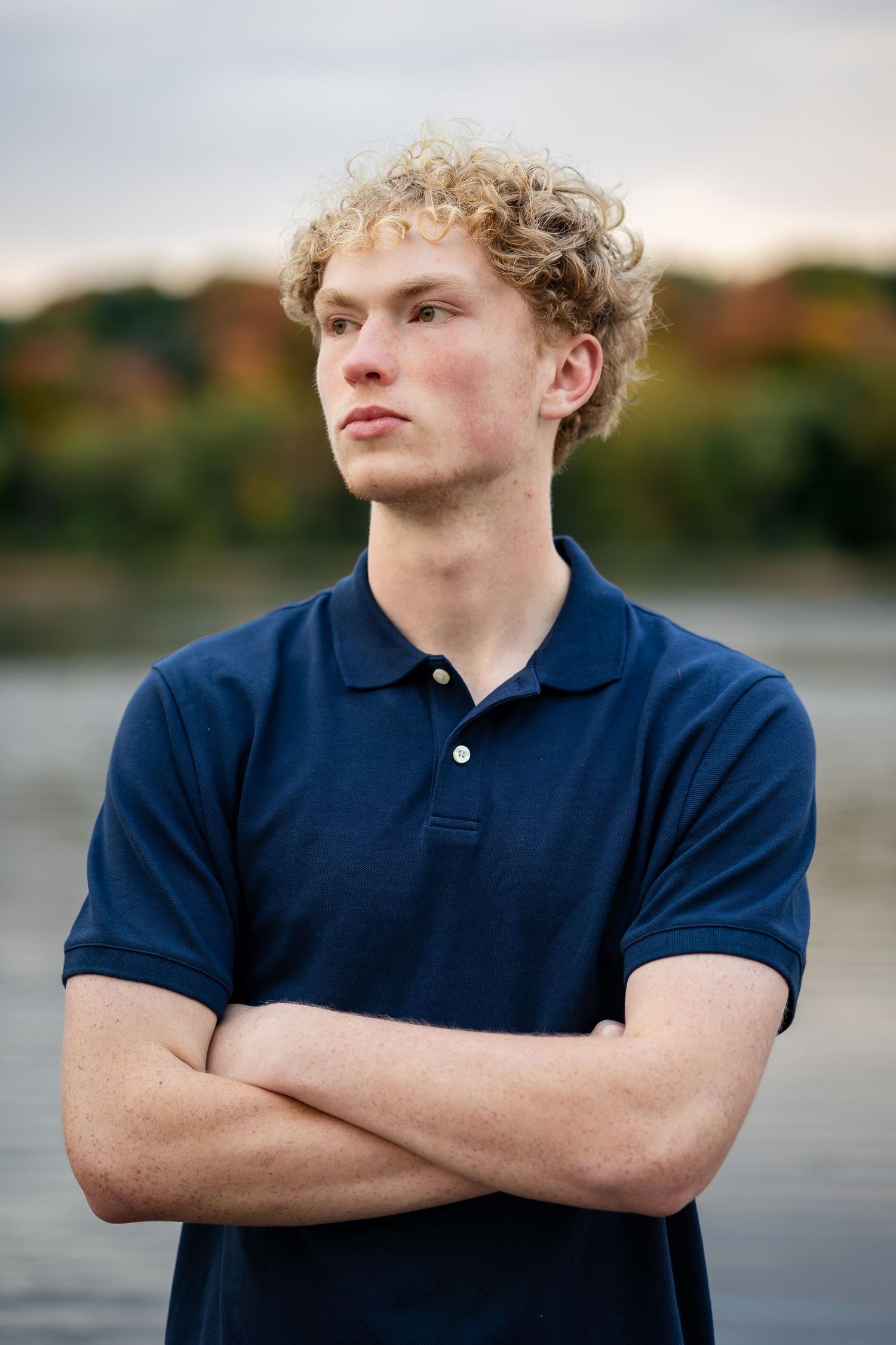 Portrait of person with curly blonde hair wearing a navy polo shirt standing with arms crossed against blurred nature background.