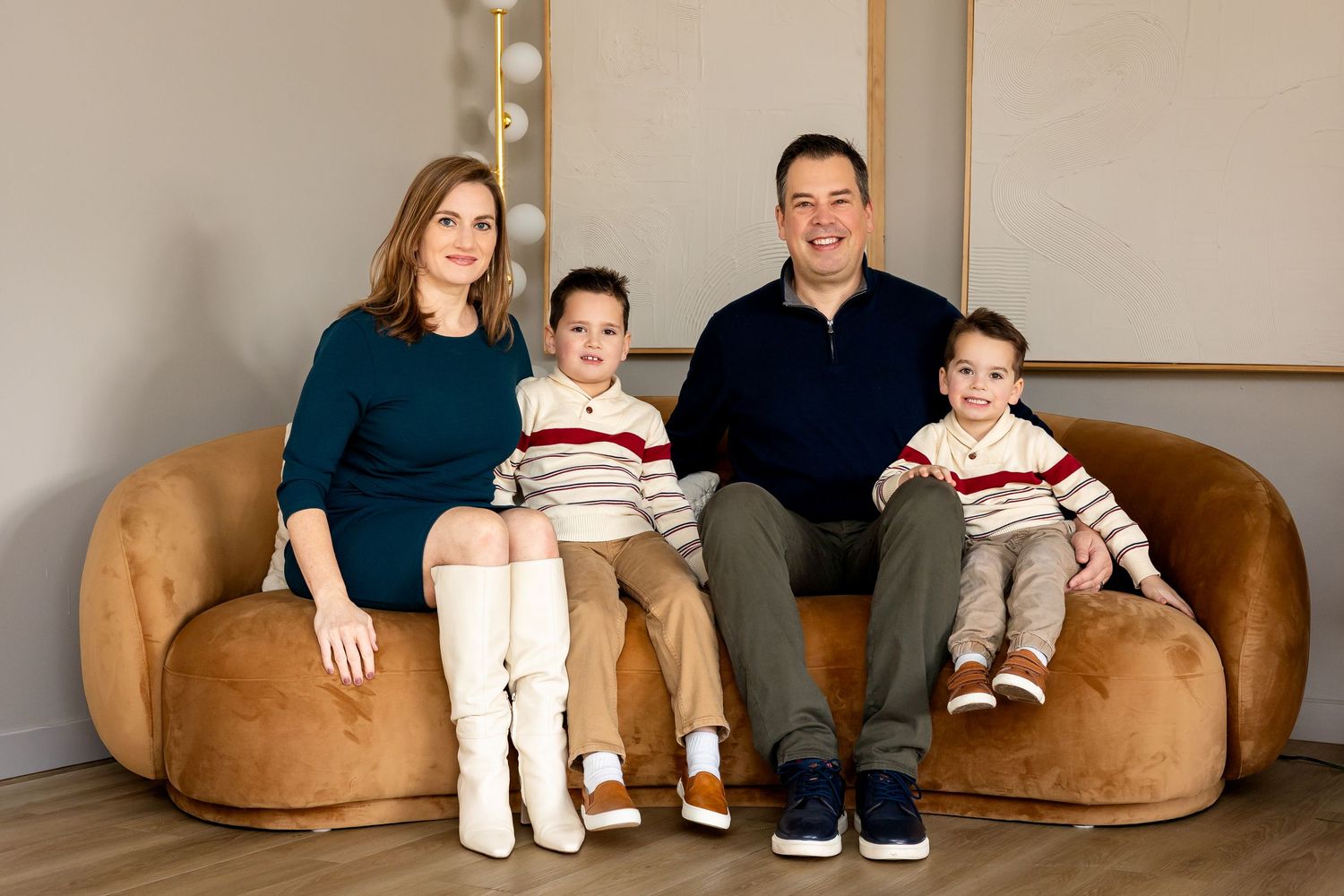 Family of four smiling on a camel sofa, two boys in striped sweaters between their parents, in a modern studio setting.
