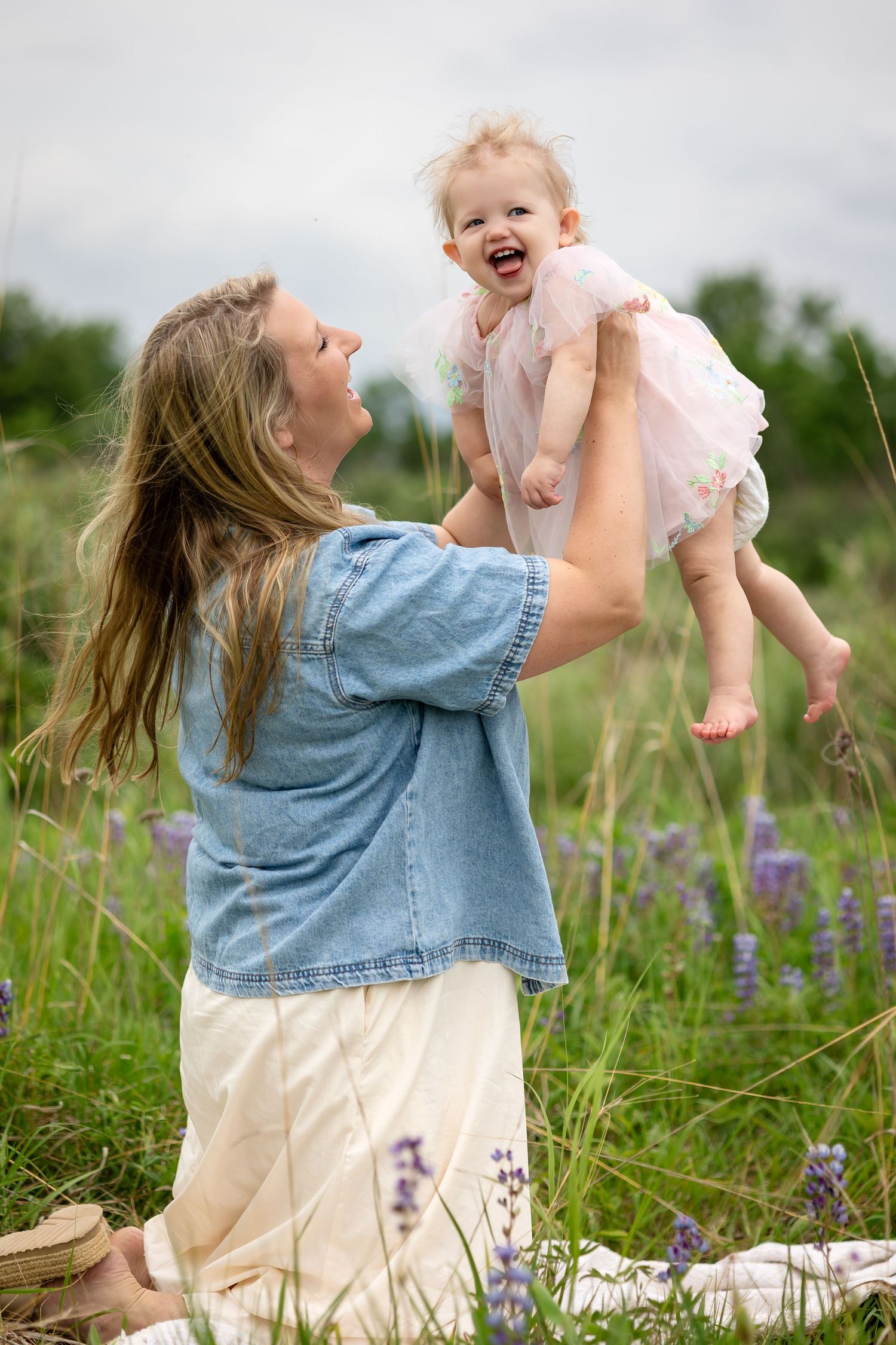 A mother lifts her laughing baby girl in a pink dress outdoors in a field of purple wildflowers.