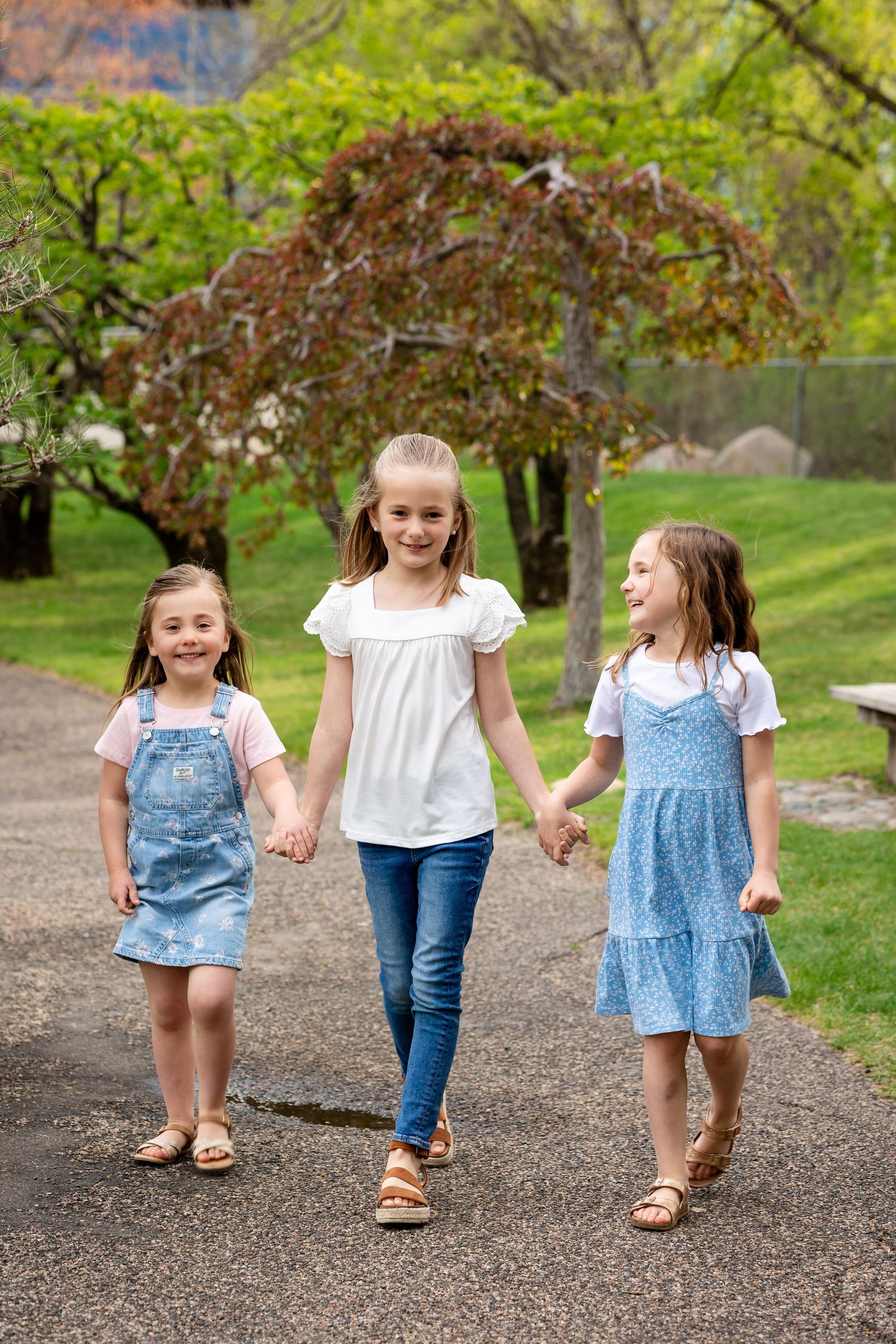 Three young girls holding hands walk together on a park path surrounded by spring trees and green grass.