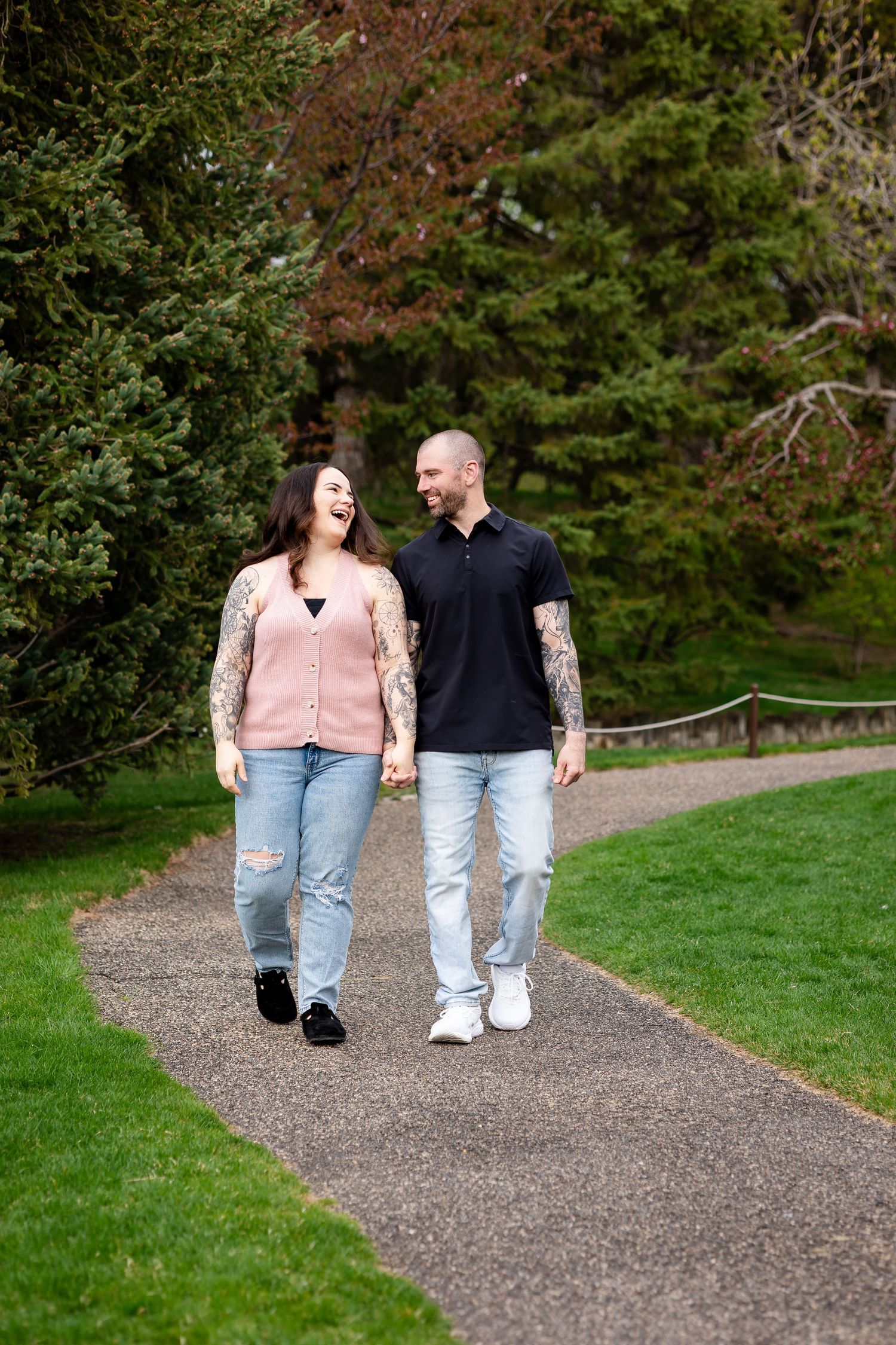 Tattooed couple laughing and holding hands while walking on a park pathway.
