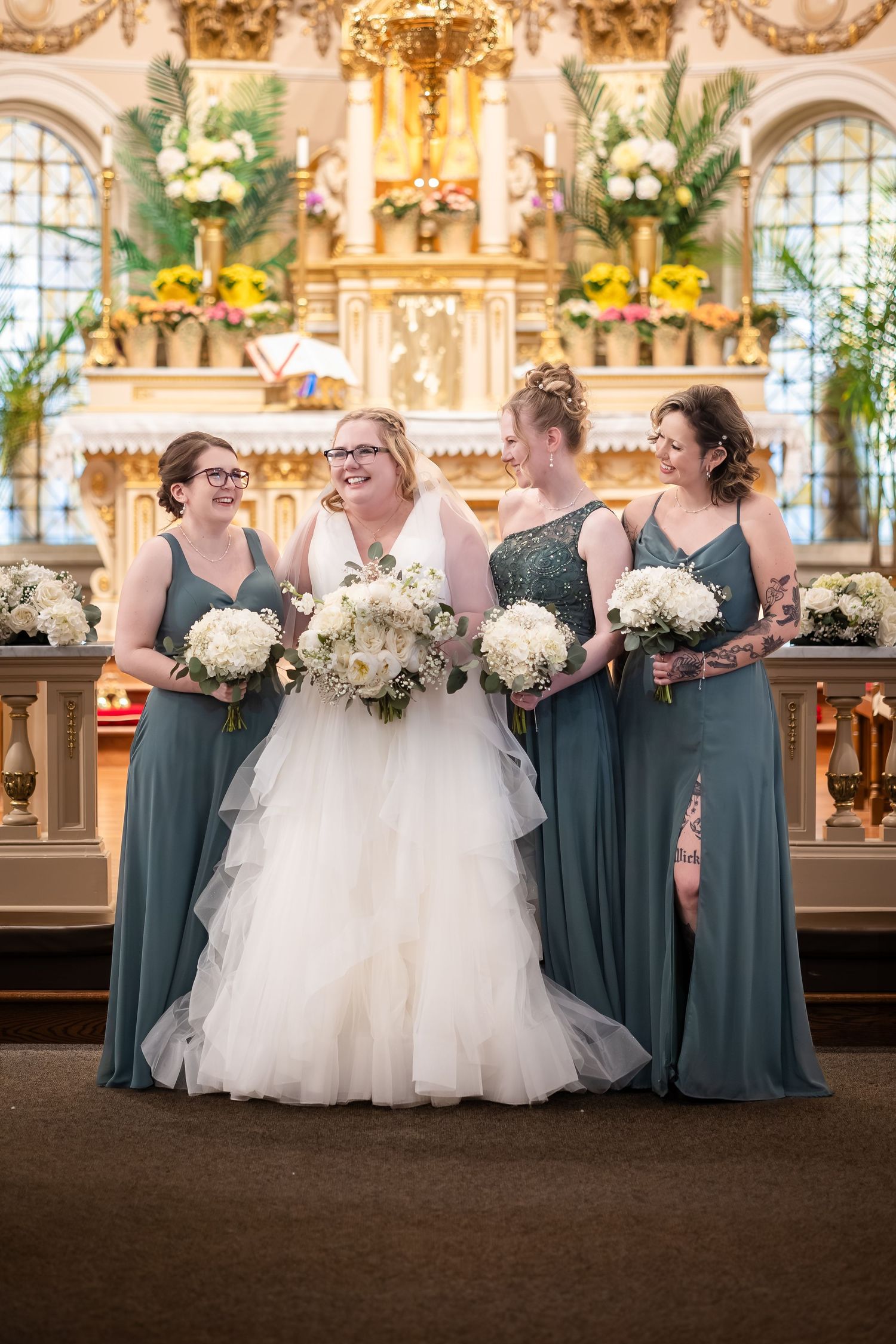 Bride in white tulle gown with three bridesmaids in sage green dresses holding white bouquets in church.