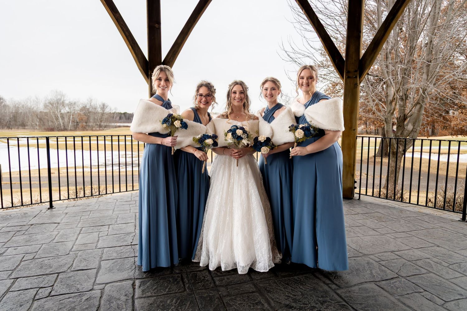 Bride in white gown poses with four bridesmaids in blue dresses and white shawls holding bouquets under a pavilion.