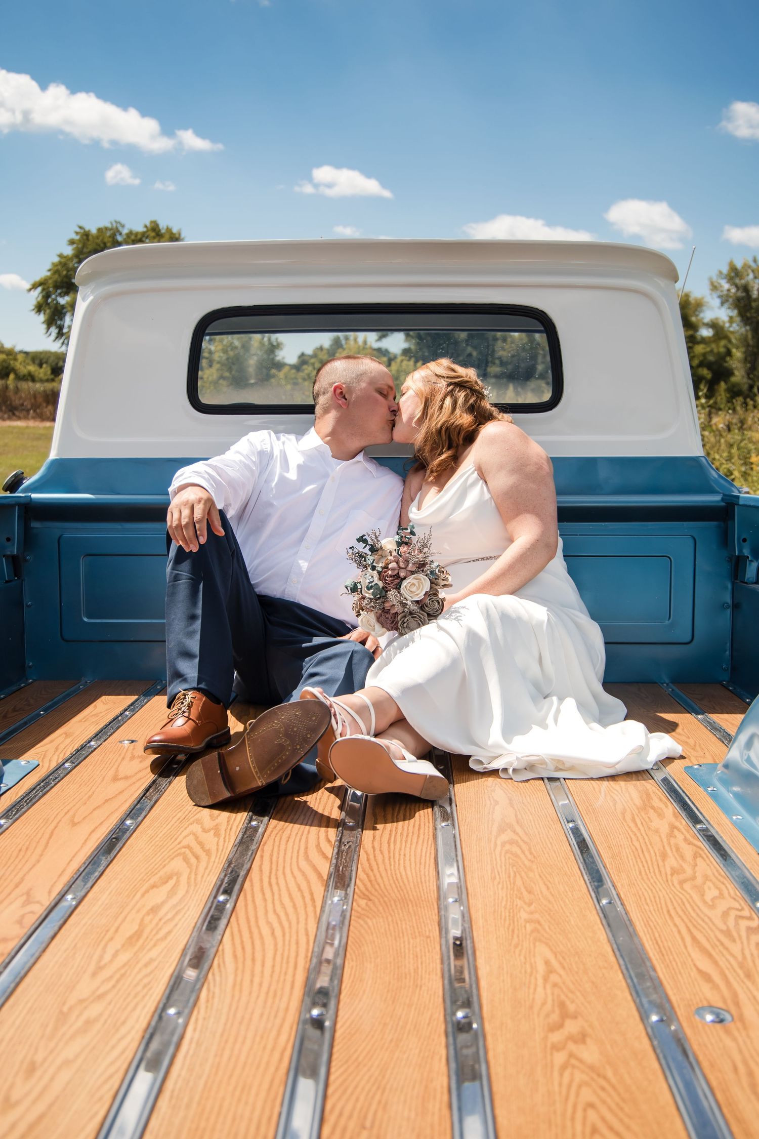 A couple in wedding attire shares romantic moments in the bed of a vintage blue pickup truck on a sunny day.