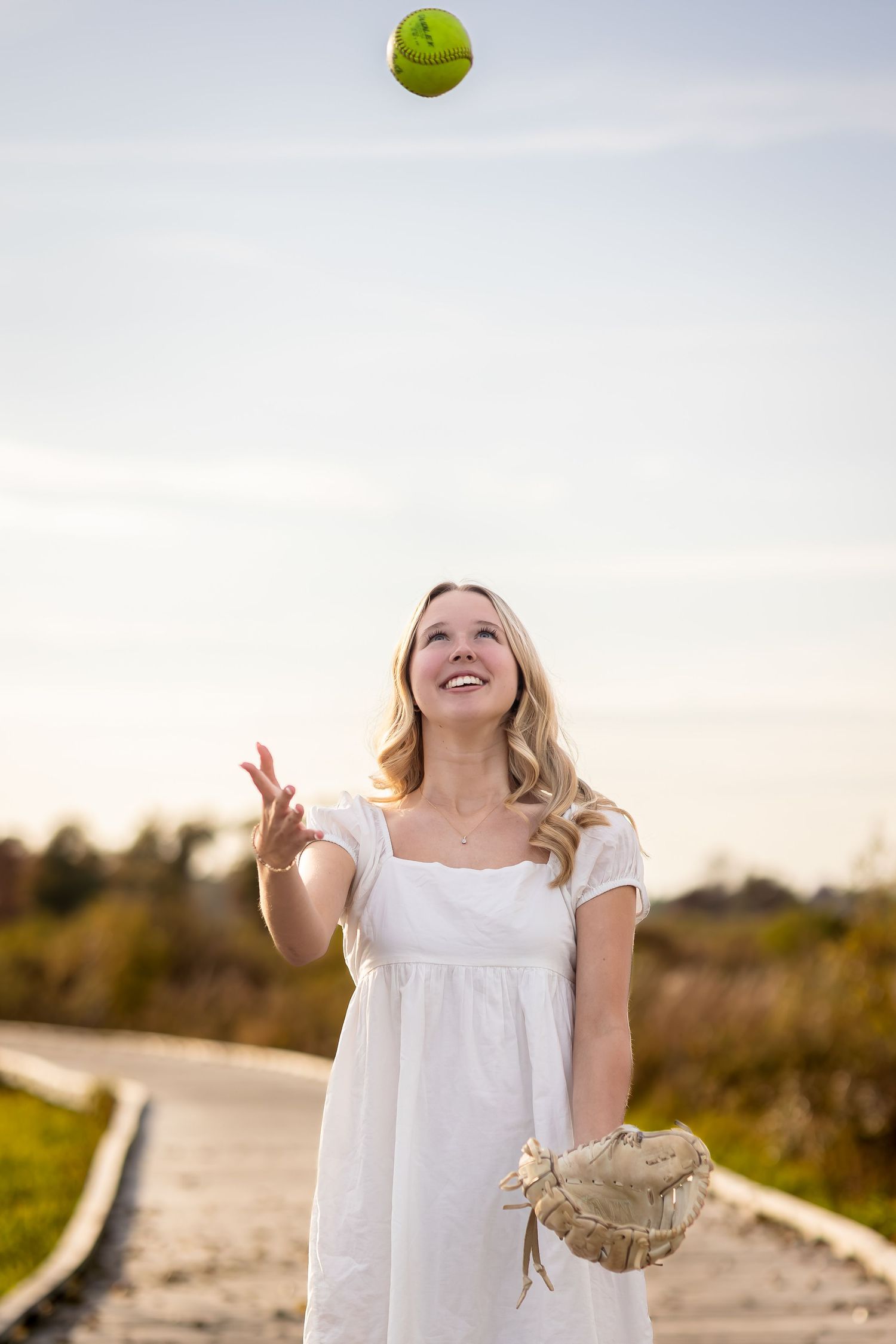 Woman in white dress plays with tennis balls on a sunny beach path during golden hour.
