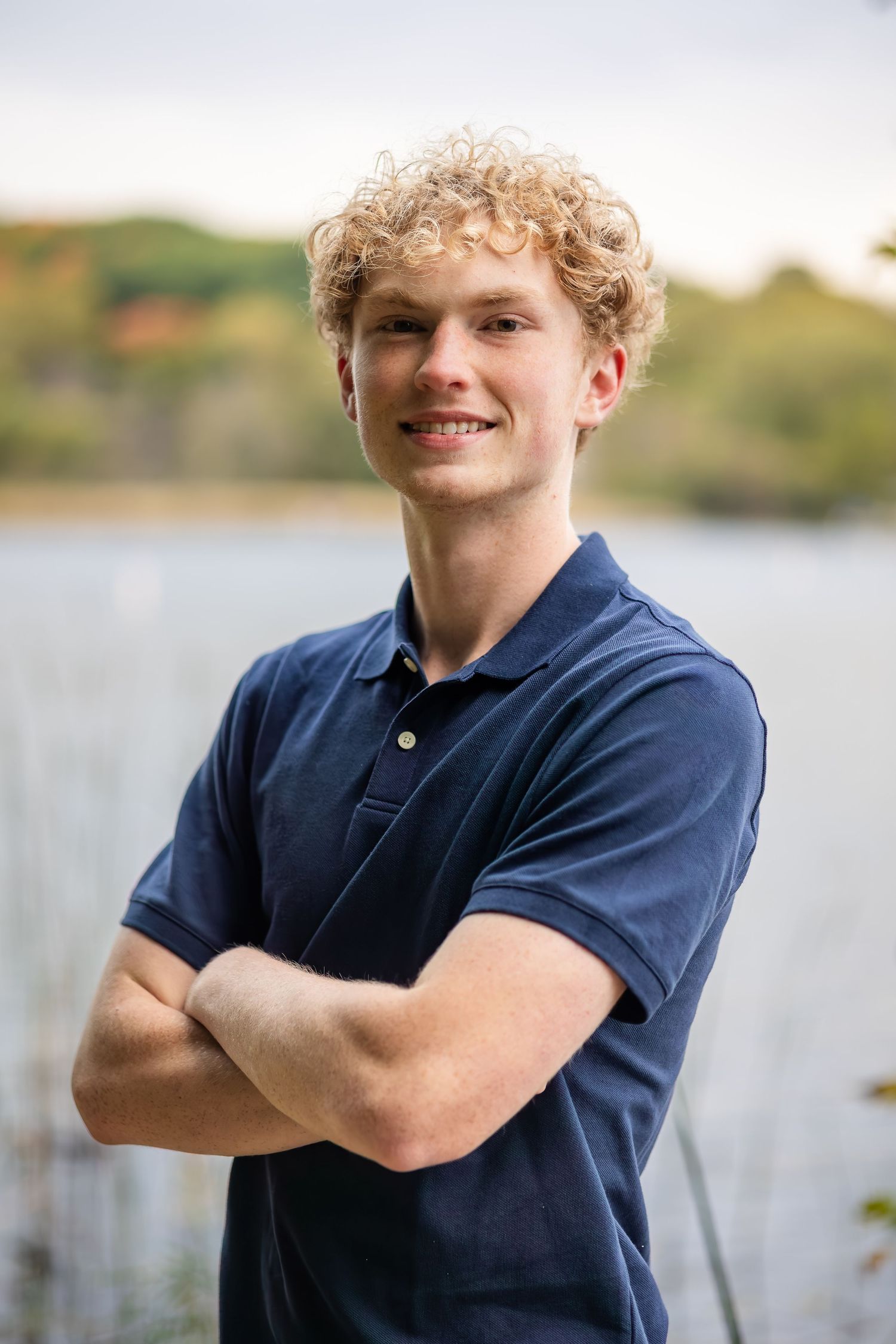 A person in a navy blue polo shirt poses outdoors by a lake with a natural background.