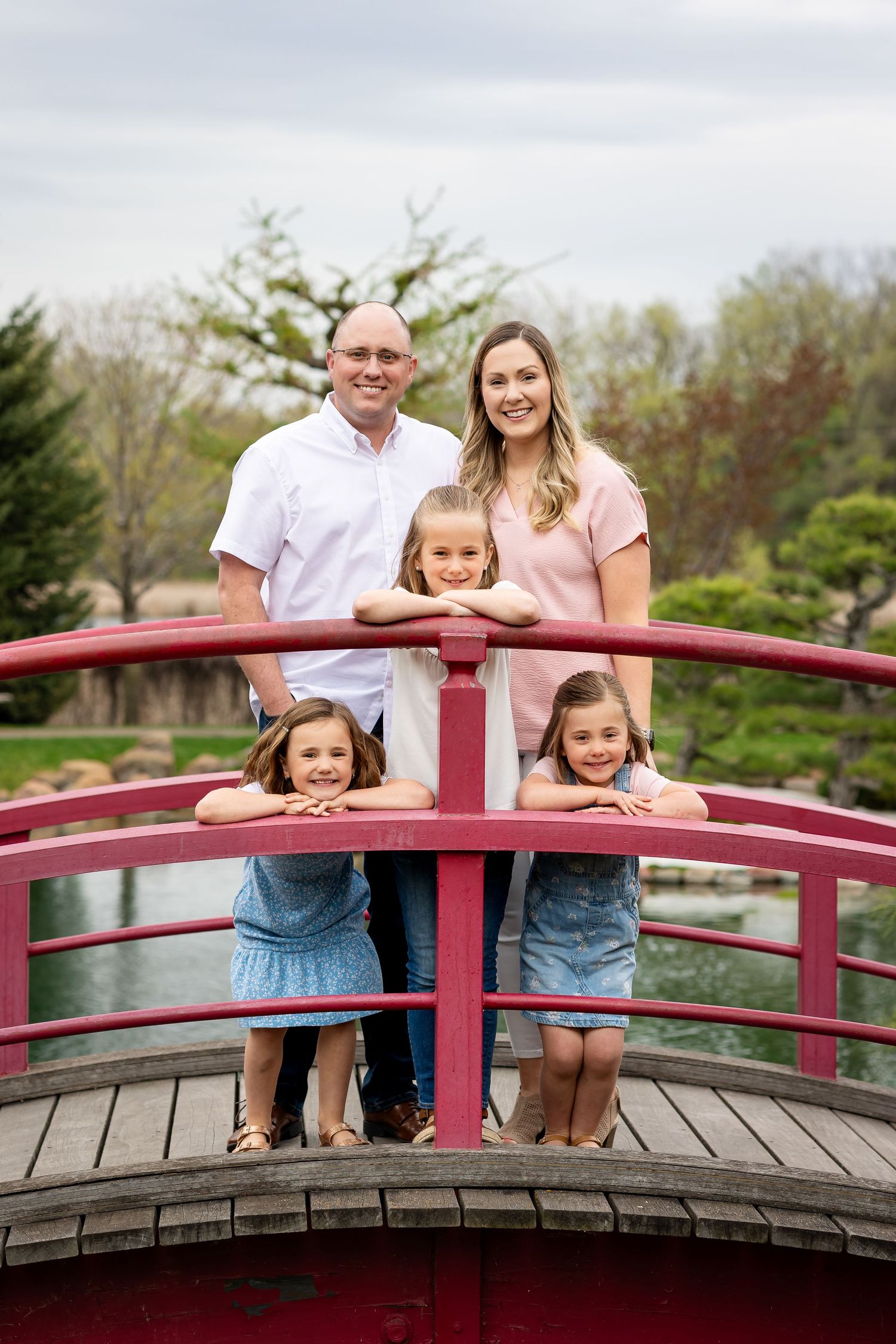 Family of five posing on a red arched bridge in a Japanese garden, parents standing behind three young girls.