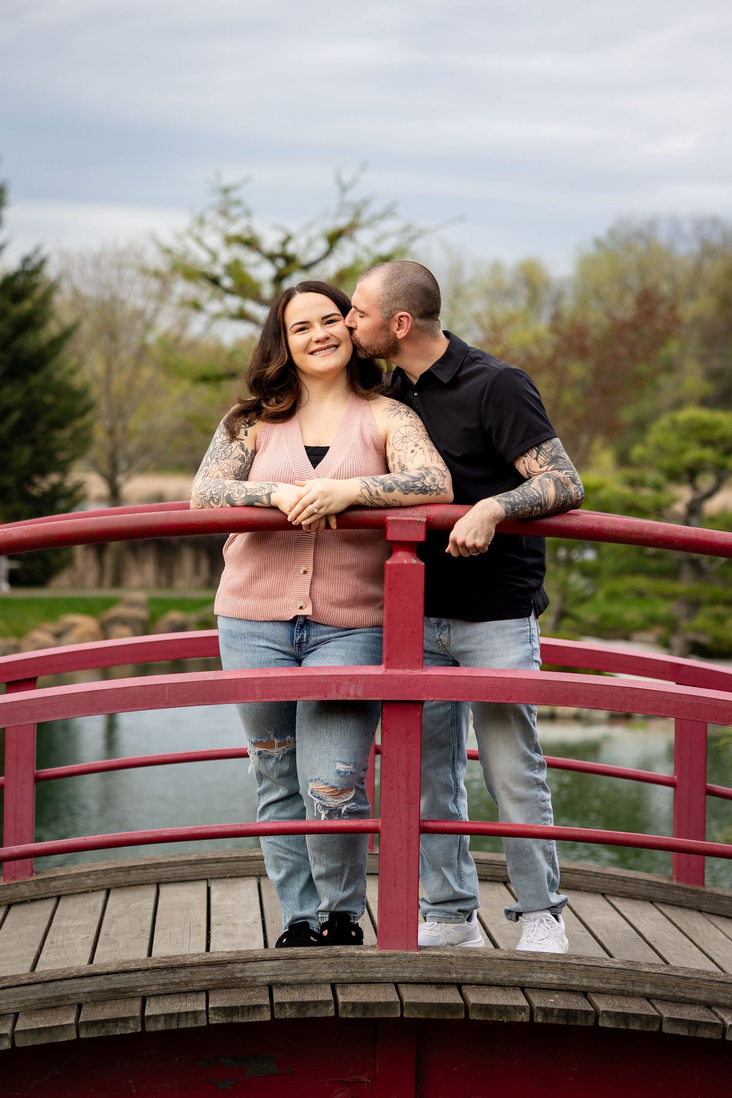 Couple with tattoos sharing a kiss on a red arched bridge in a Japanese garden park.
