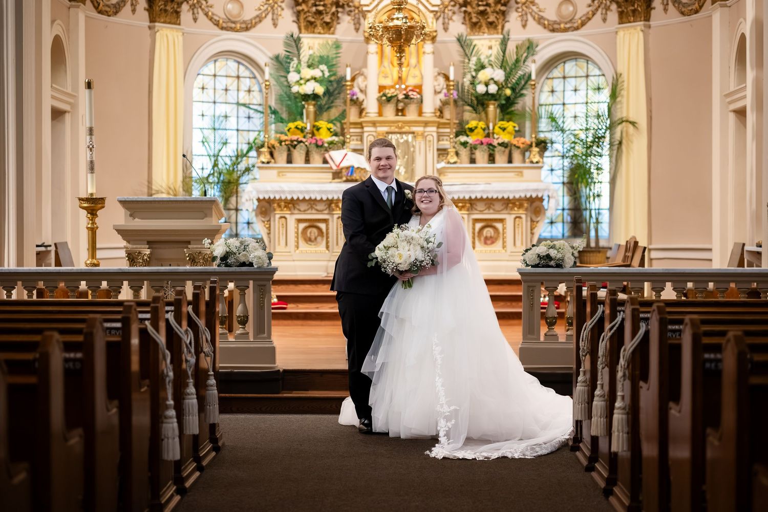 Bride and groom pose together at church altar in elegant wedding attire, surrounded by floral arrangements and wooden pews.
