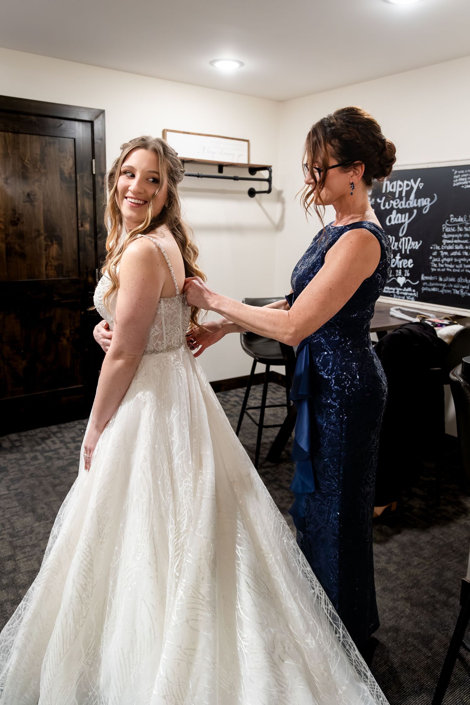 Bride in white lace gown smiling as woman in navy dress buttons her wedding dress in a bridal suite.