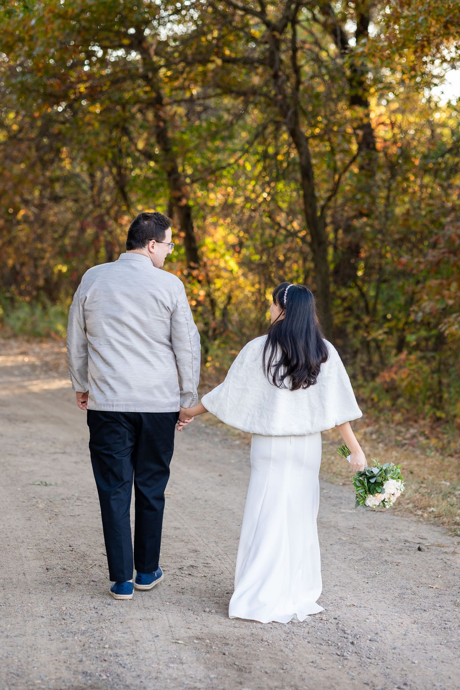 A couple walks hand in hand on a dirt path through autumn woods.