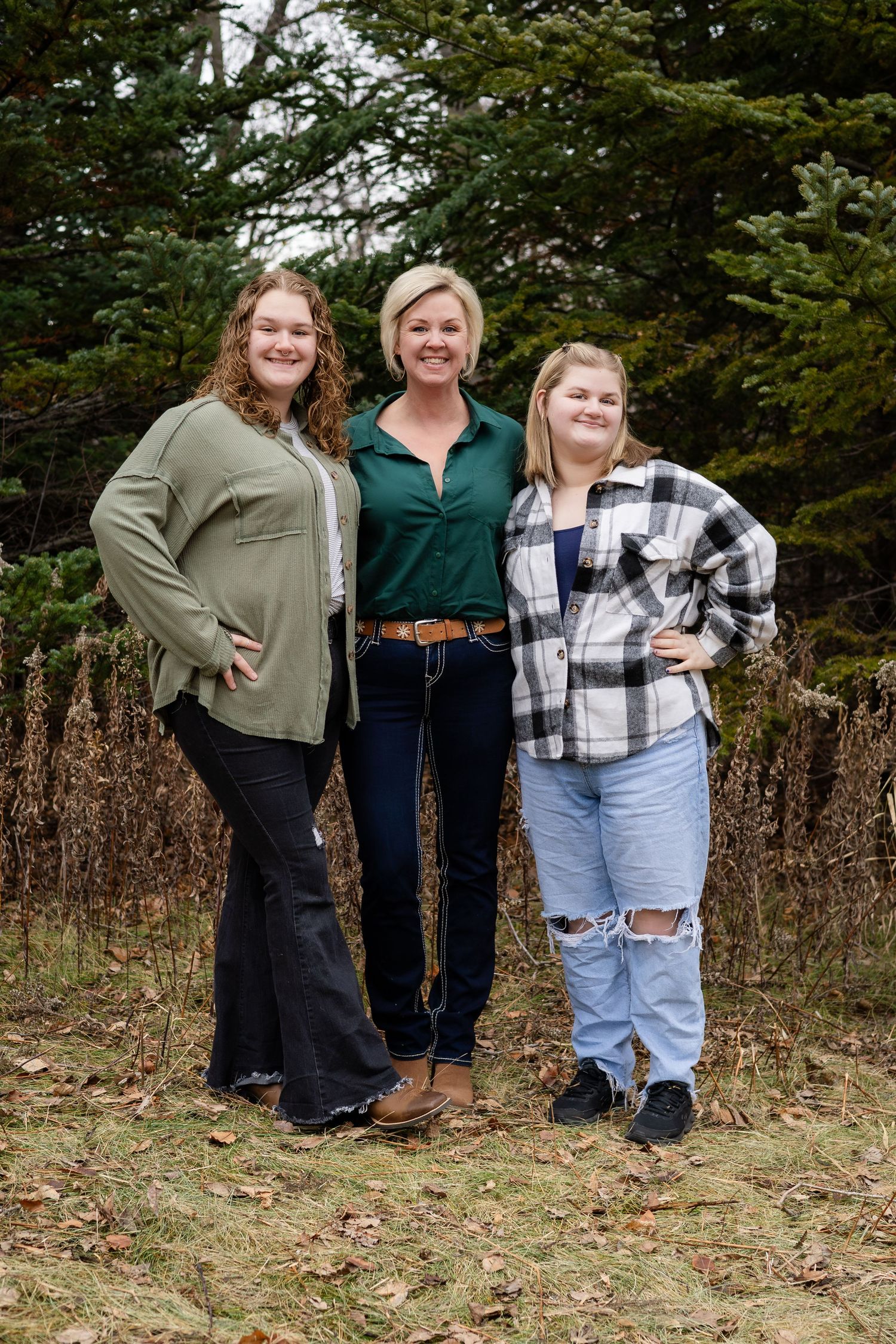 Three people in casual outfits stand together outdoors against a natural backdrop of trees and dried foliage.