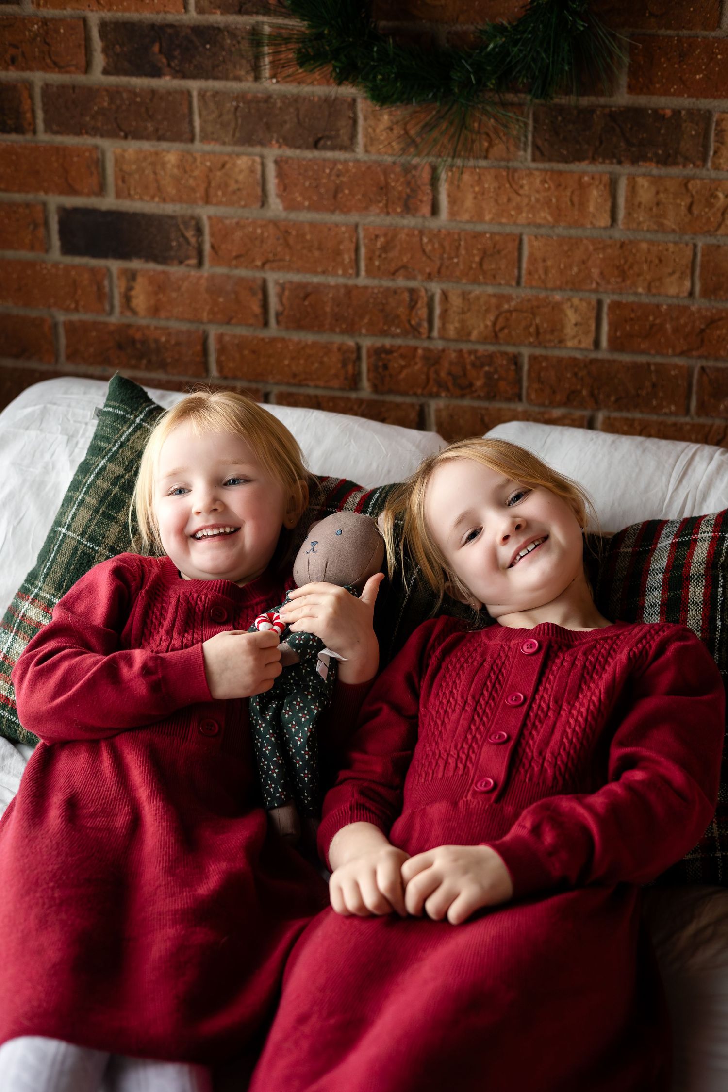 Children in red sweaters lying together on white cushions against brick wall with festive decor.