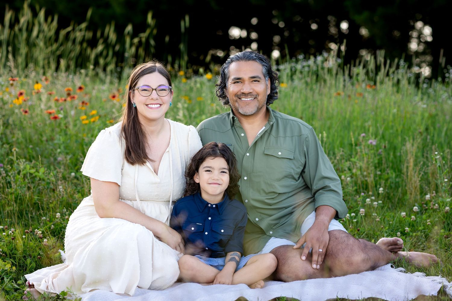 A family sits together on a white blanket in a wildflower meadow with orange poppies and tall grasses on a sunny day.