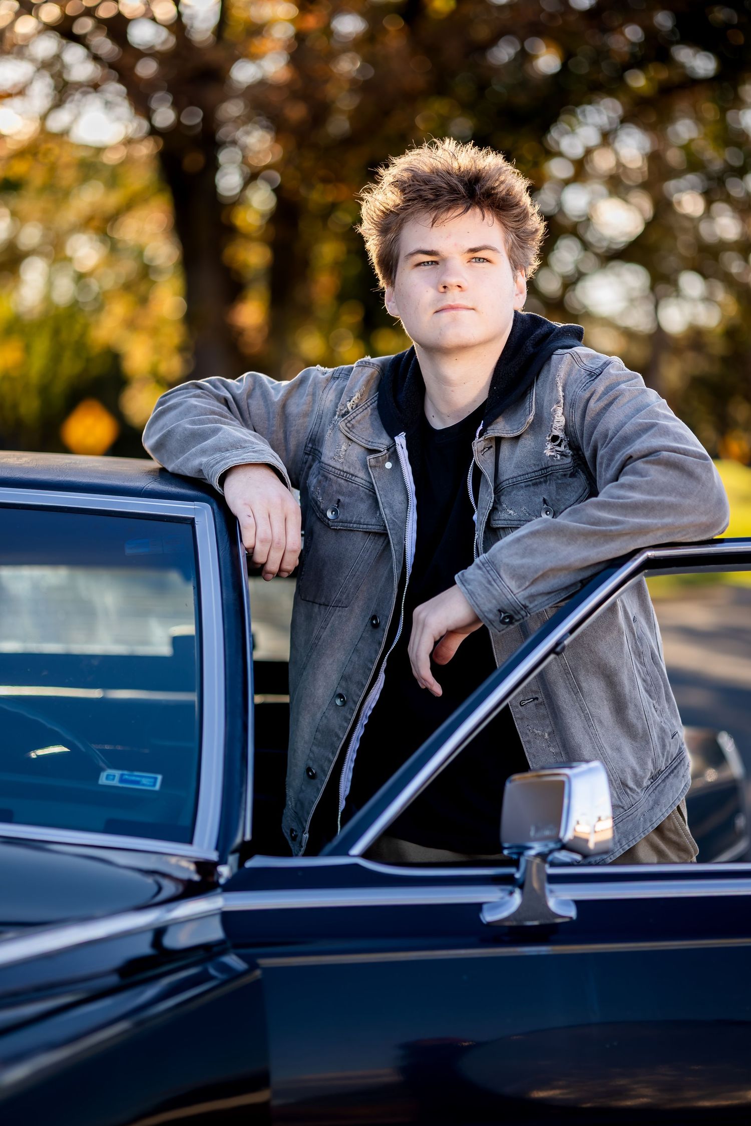 A young person in a gray denim jacket leans against a classic black car during golden hour with autumn trees in background.