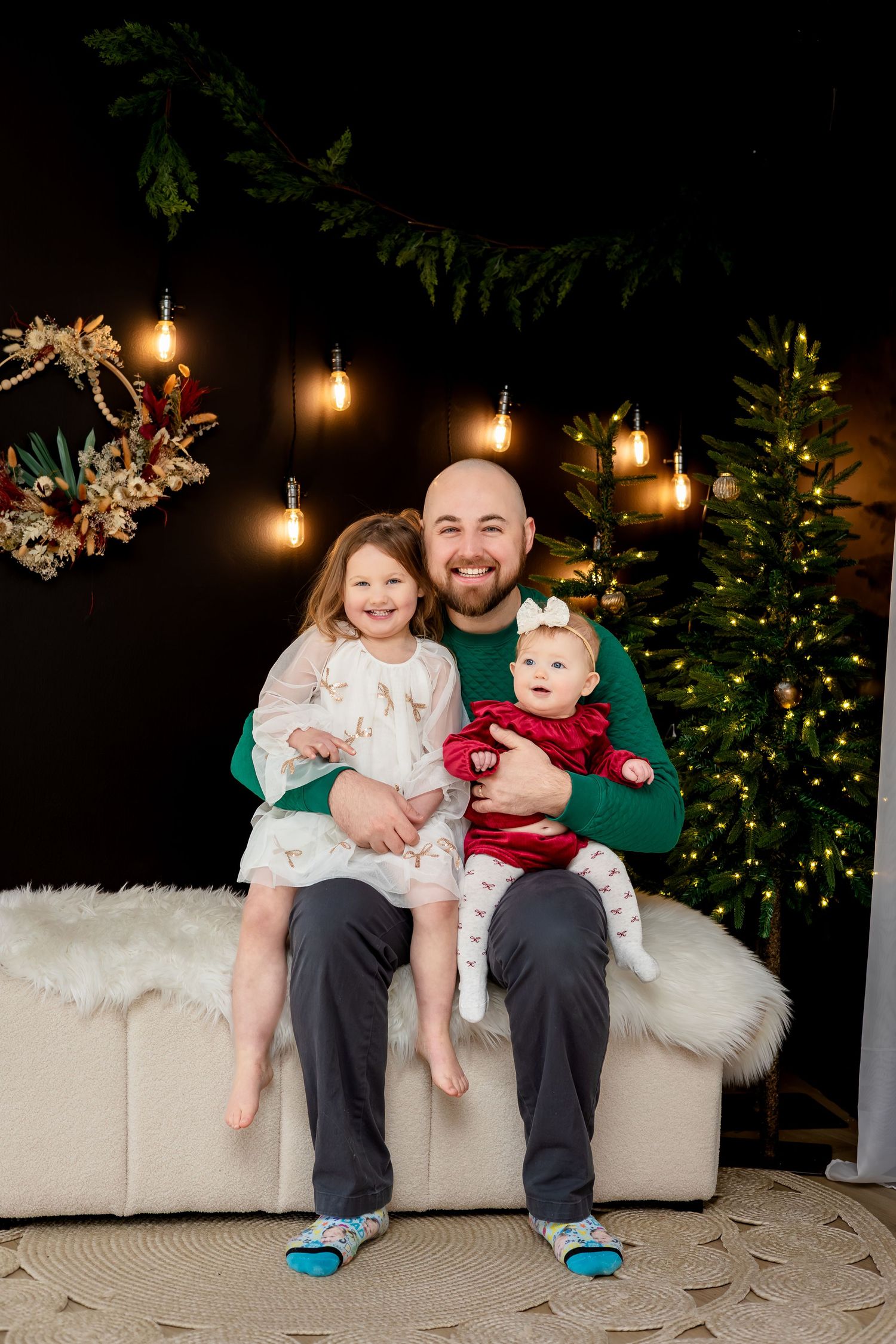 Smiling bald father sits on white couch holding two young daughters in front of Christmas tree with festive holiday lights.