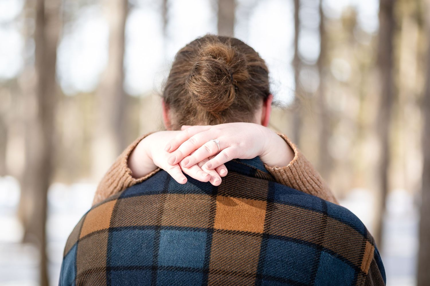Couple embracing outdoors in winter forest, woman's hand resting on man's shoulder wearing plaid scarf.