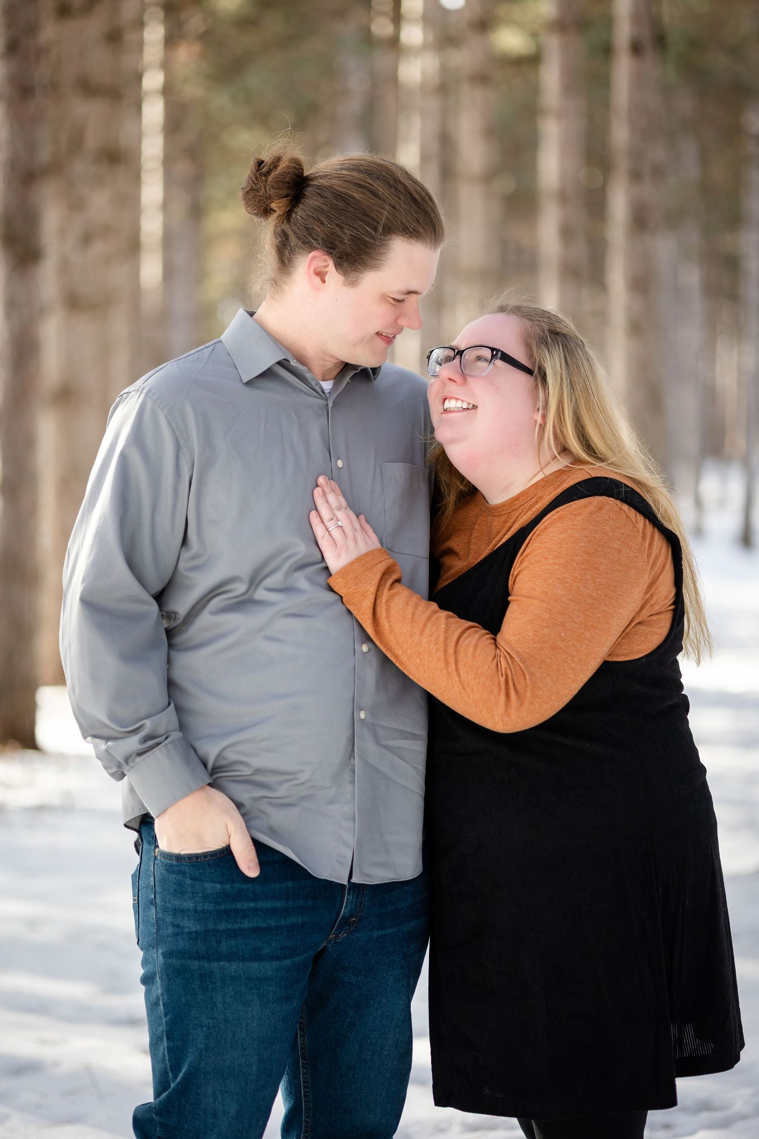 Couple sharing a romantic moment in a snowy forest, woman in orange and black smiling up at man in gray shirt.