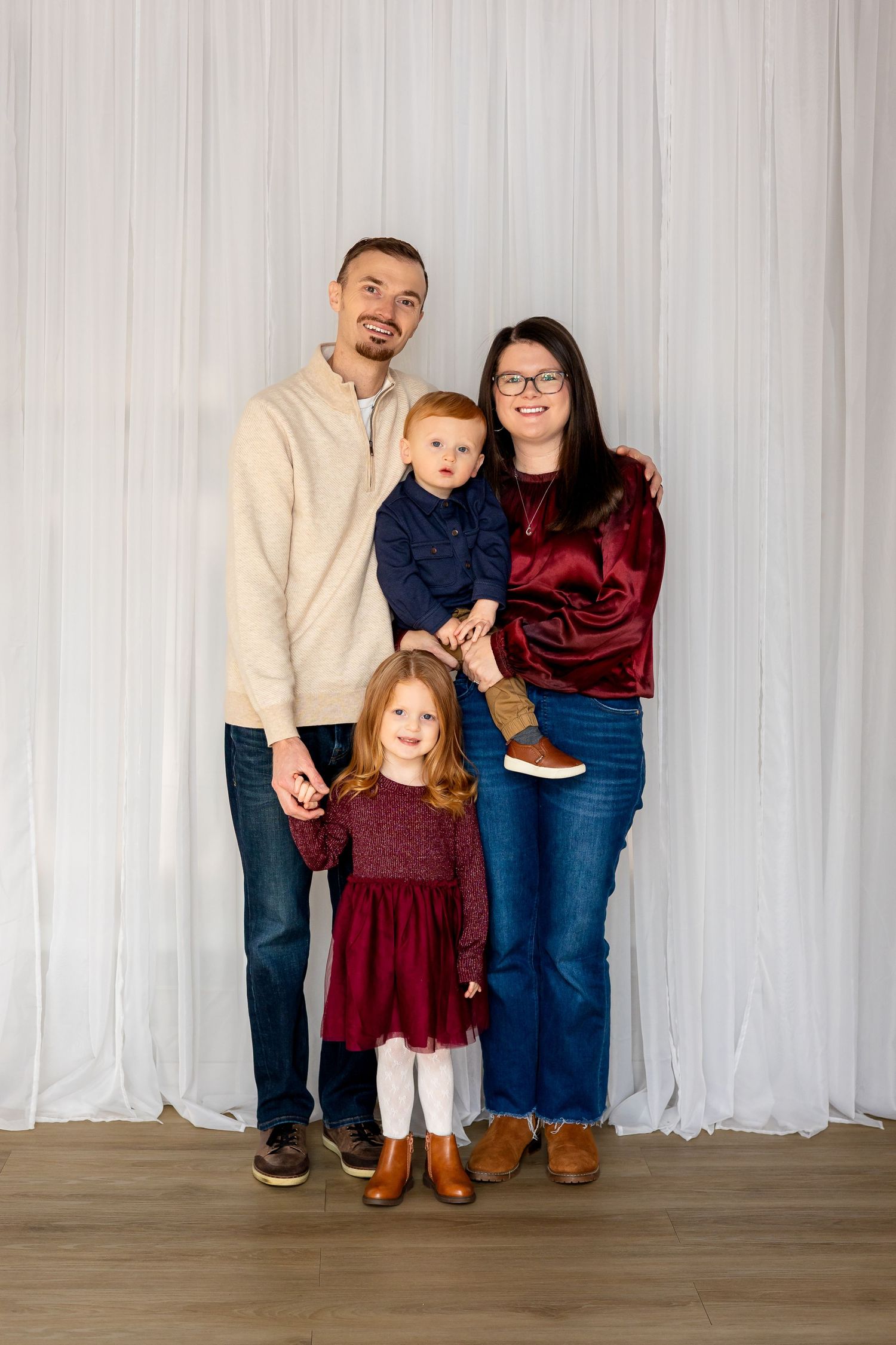 Family of four posing for a portrait, parents holding two young children, dressed in coordinating burgundy and navy outfits.