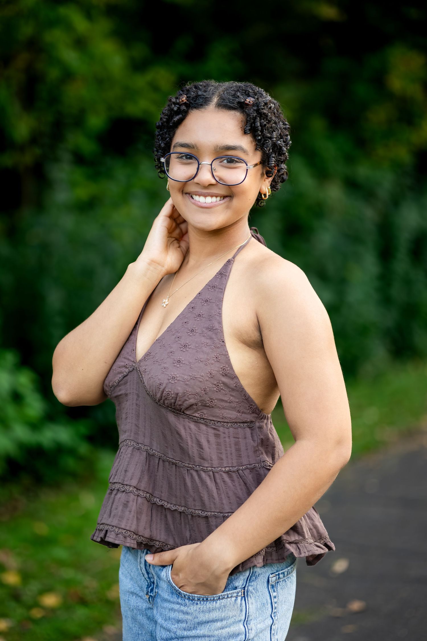 A person wearing glasses and a brown sleeveless top poses outdoors on a path with greenery in the background.