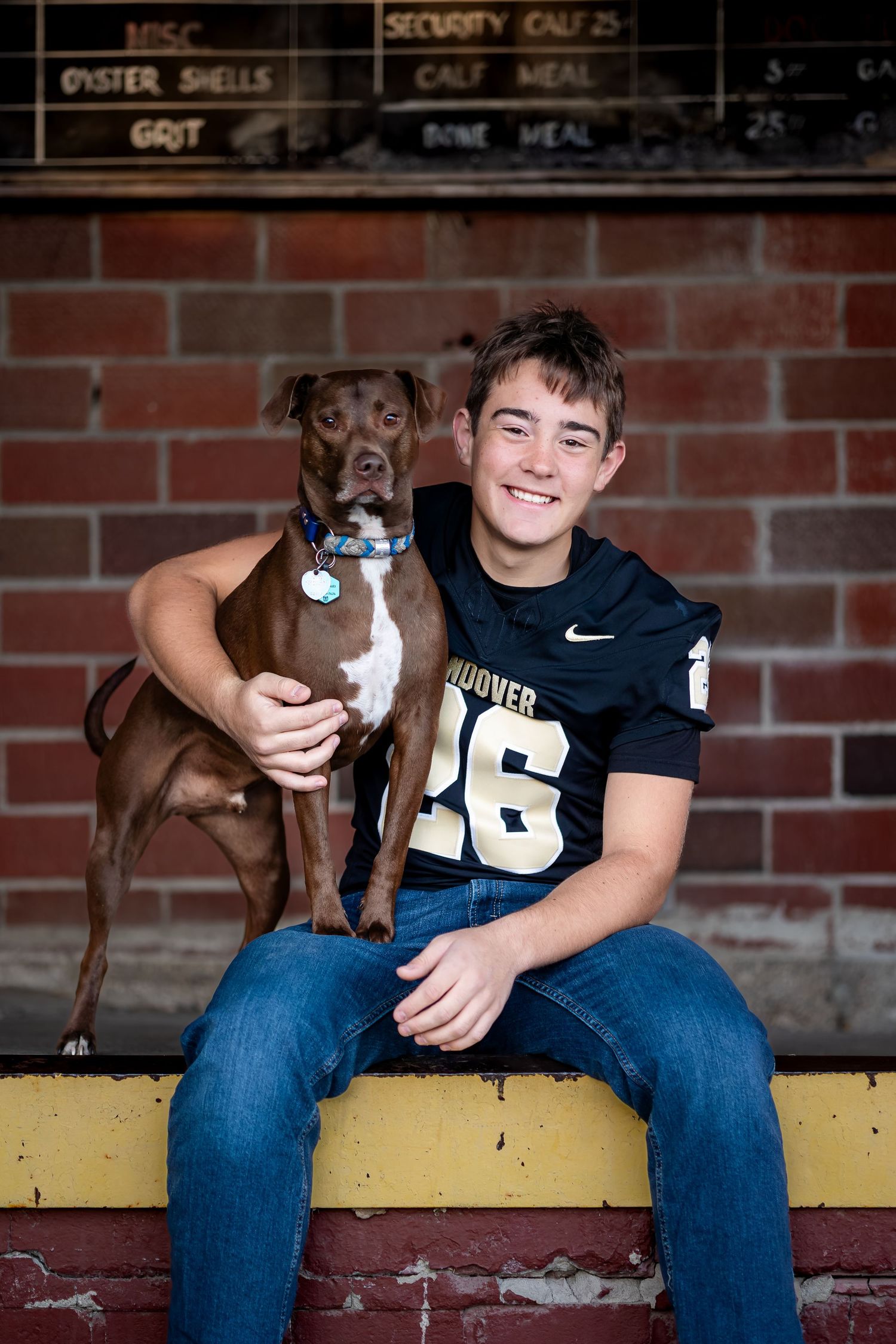 A brown dog and athlete in jersey number 26 sit together on a yellow bench against a brick wall.