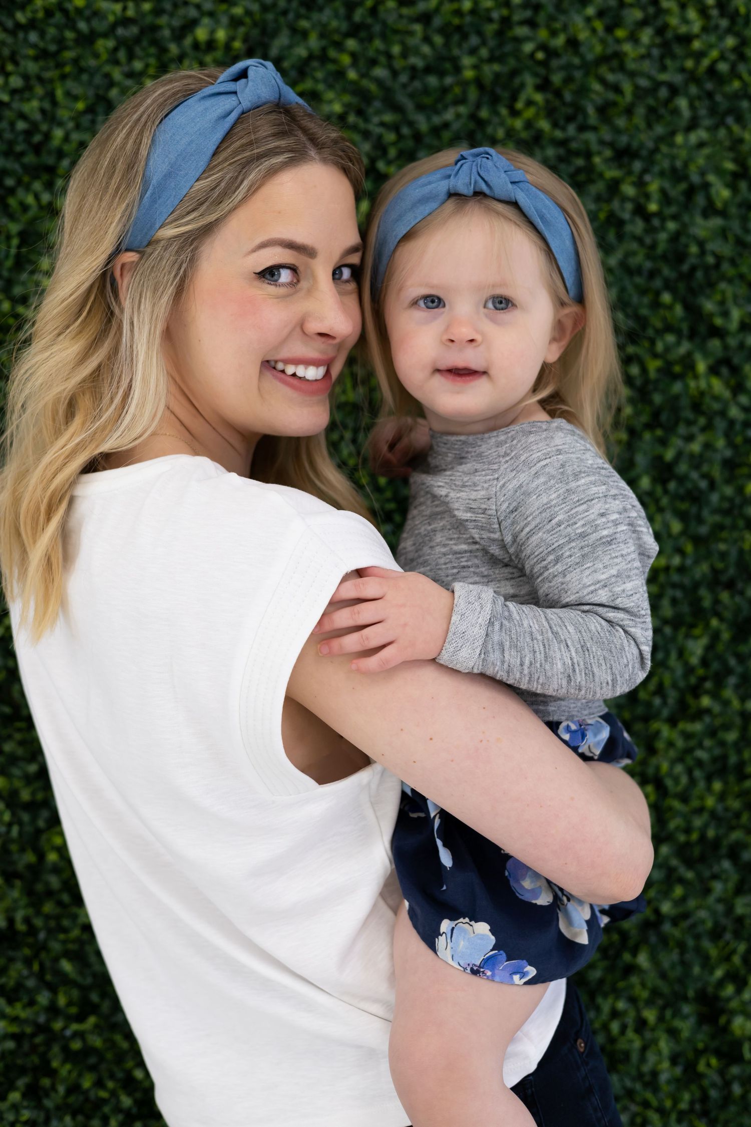 A cheerful portrait against a green hedge backdrop featuring matching blue headbands and floral dress.