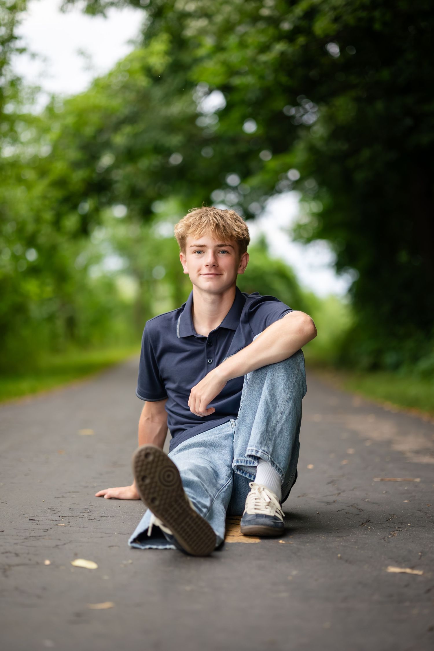 Person sits casually on ground of tree-lined pathway in casual attire.