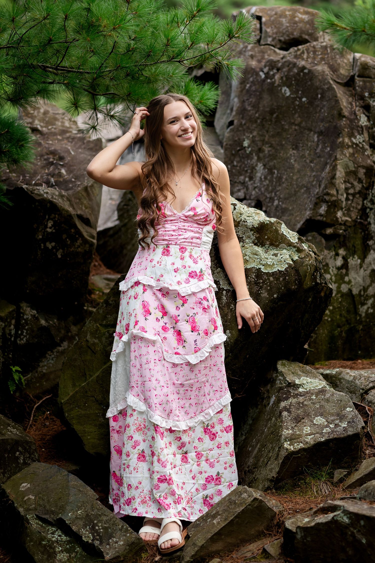 Woman in layered pink floral dress stands among rocky terrain and pine trees.