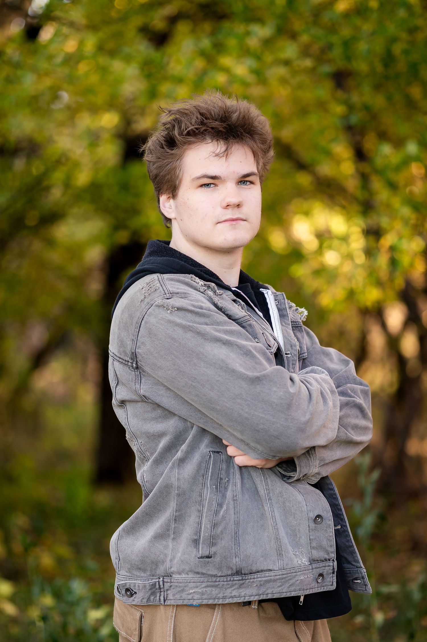A person in a gray denim jacket poses outdoors with crossed arms against a blurred autumn background.