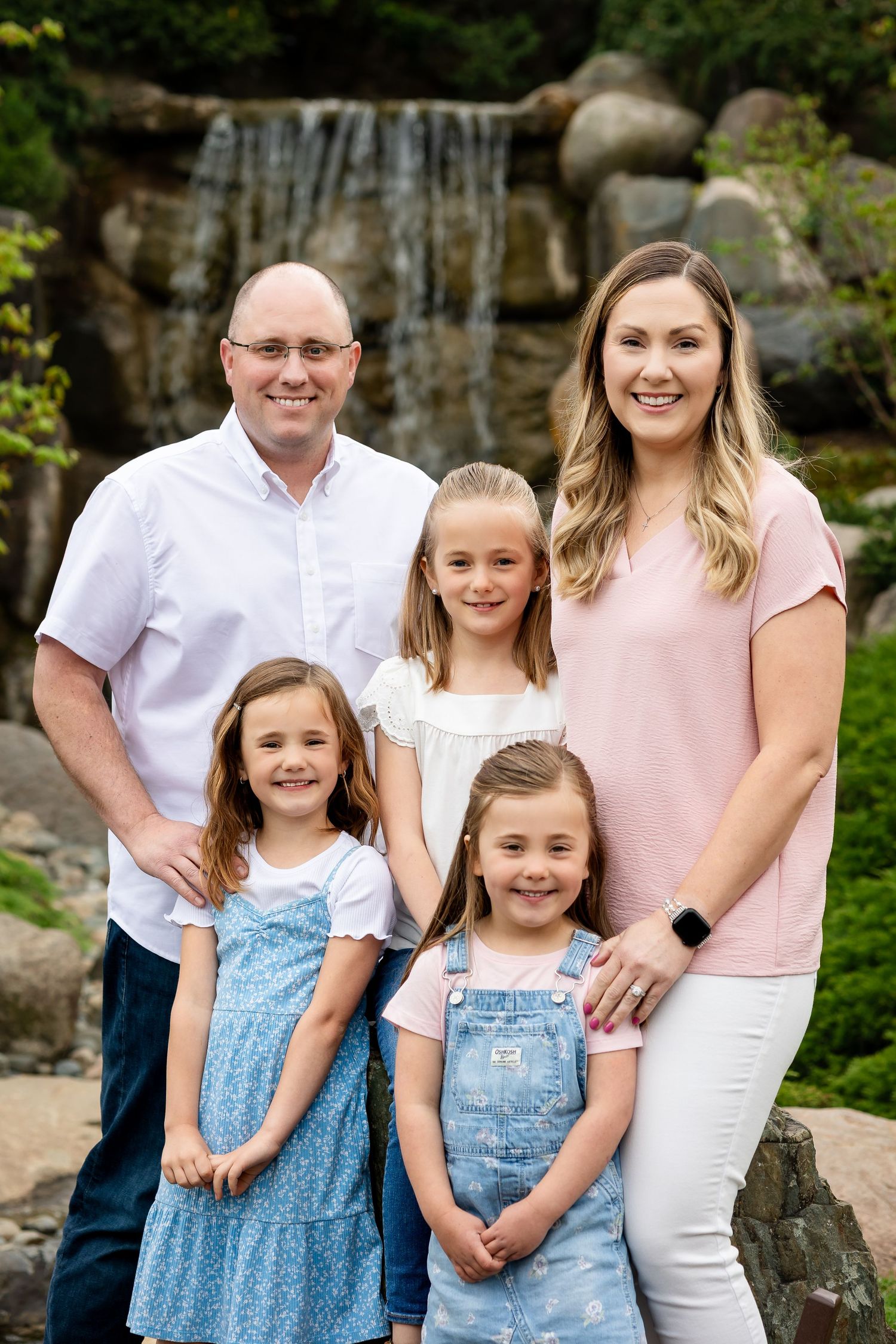 Happy family of five posing in front of a scenic waterfall backdrop in a lush green garden.