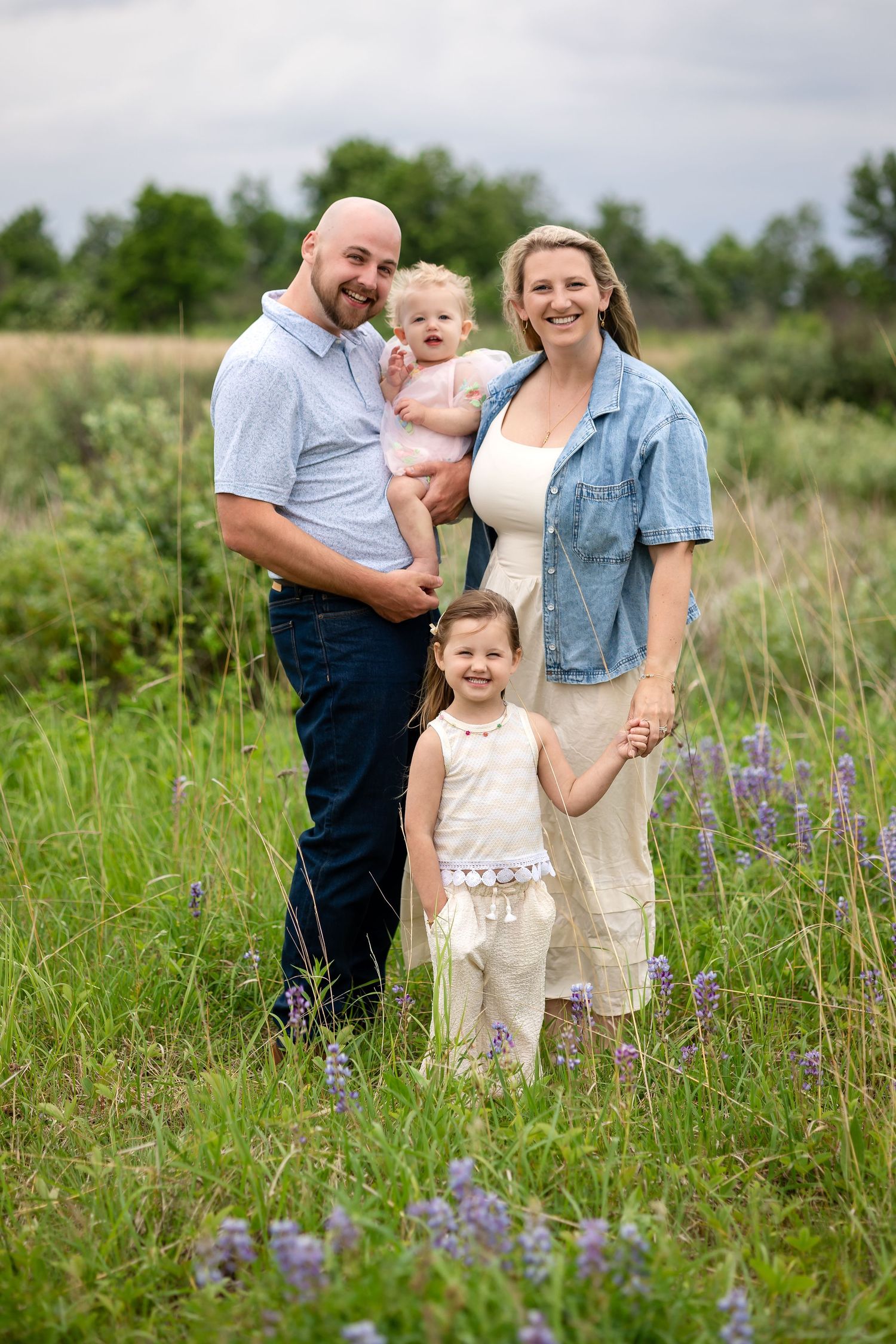 Family of four posing in a wildflower meadow, parents holding baby and toddler girl smiling outdoors.