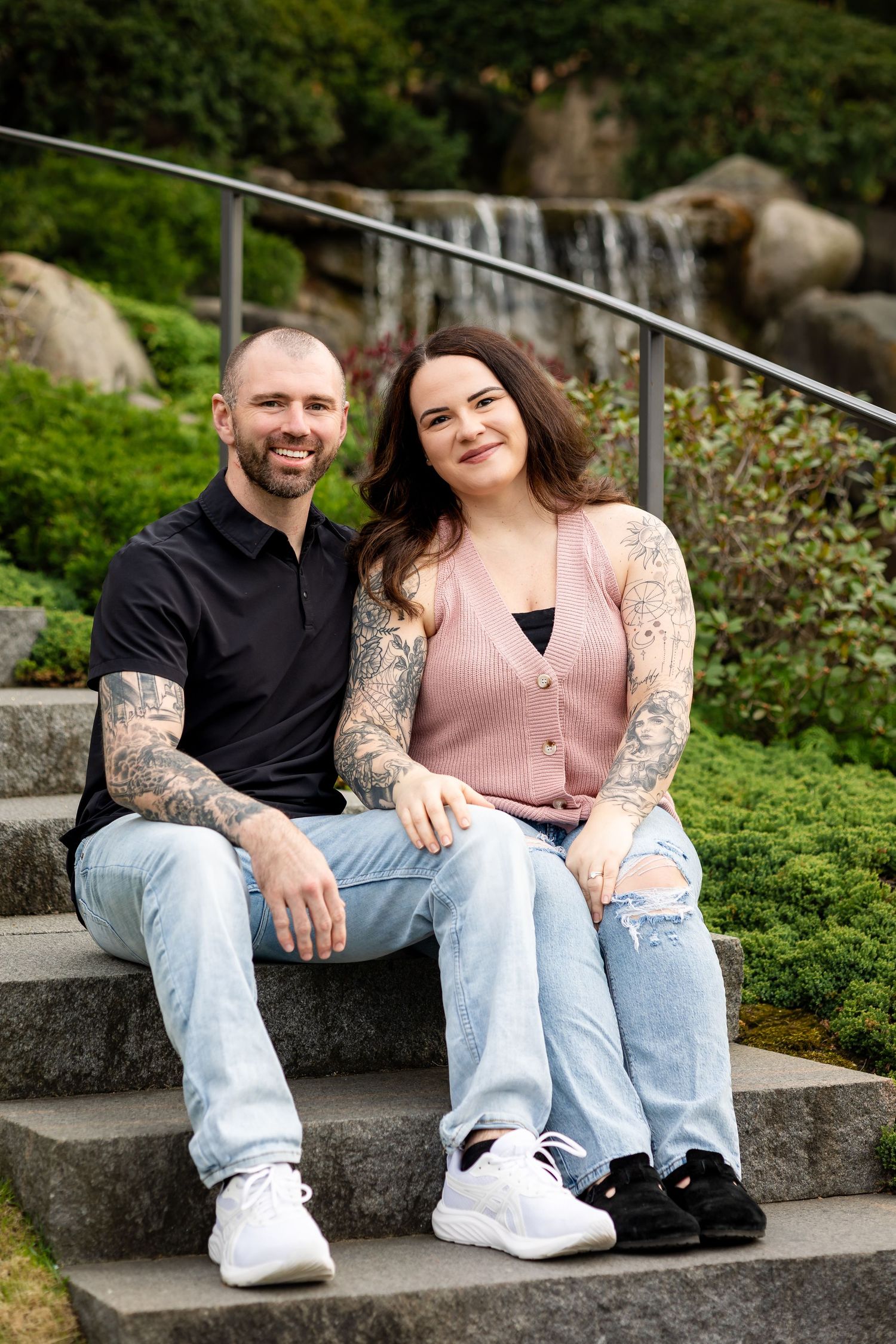 Tattooed couple sitting on stone steps in front of a waterfall and lush greenery, smiling at the camera.