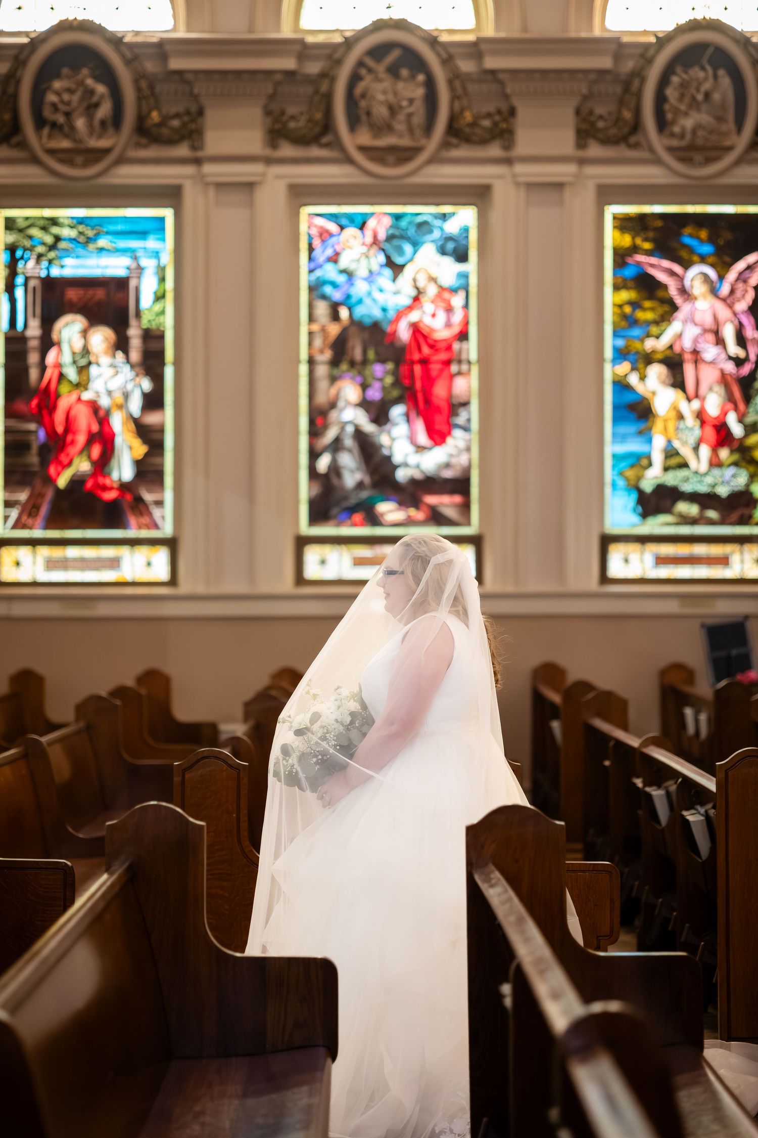 Bride in white gown and veil kneeling in church pew before colorful stained glass windows.