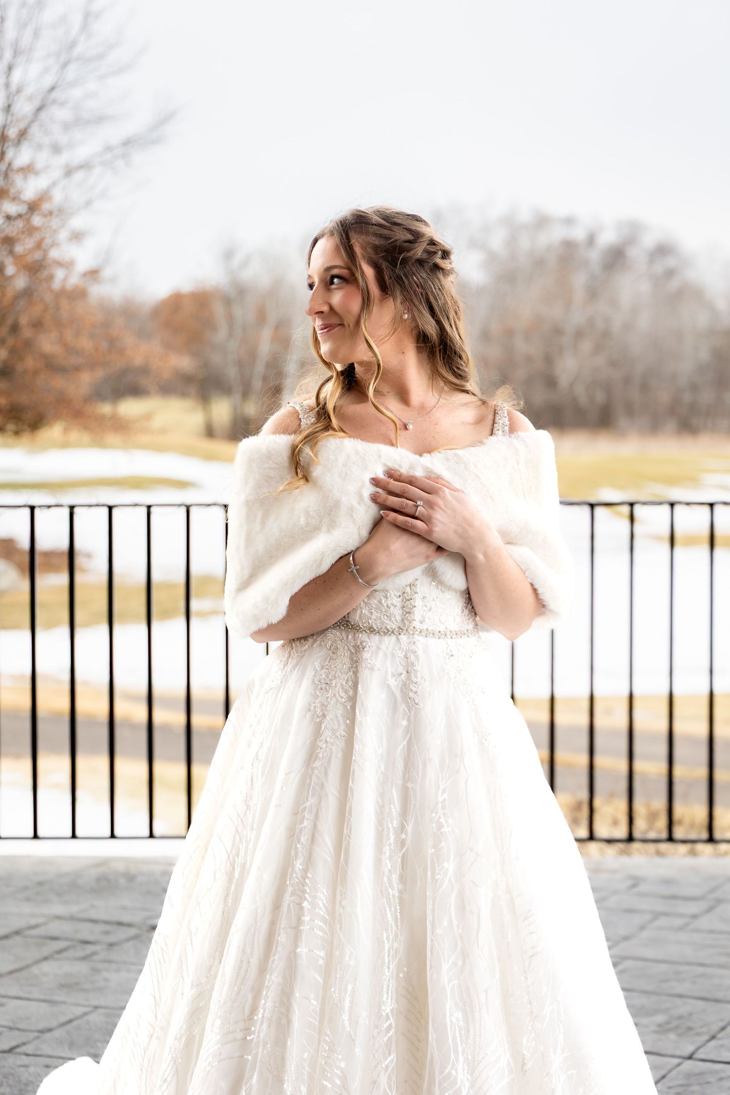 Bride in lace ball gown and white fur wrap poses on outdoor terrace in winter setting.