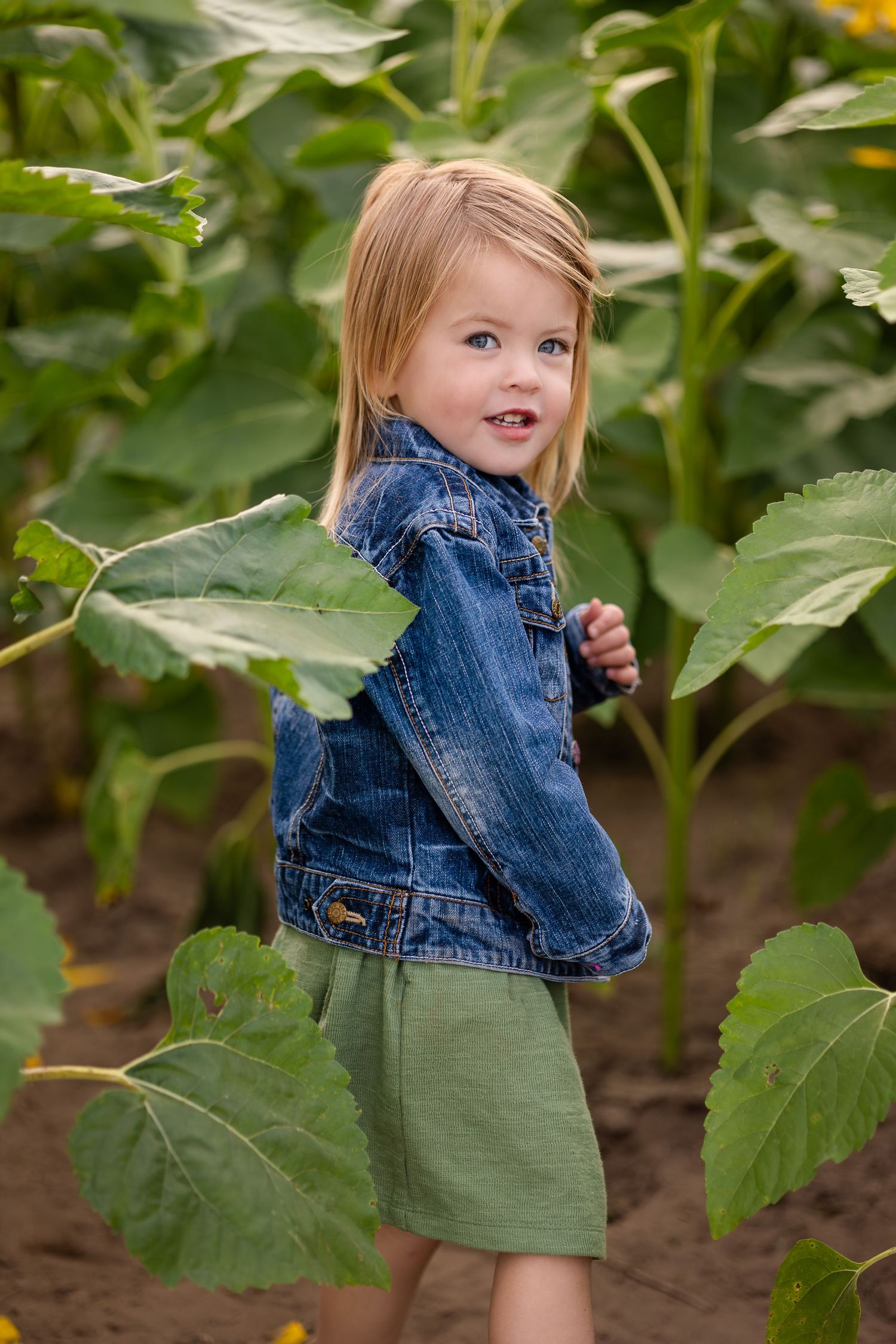 Child in denim jacket and green dress exploring among garden plants.