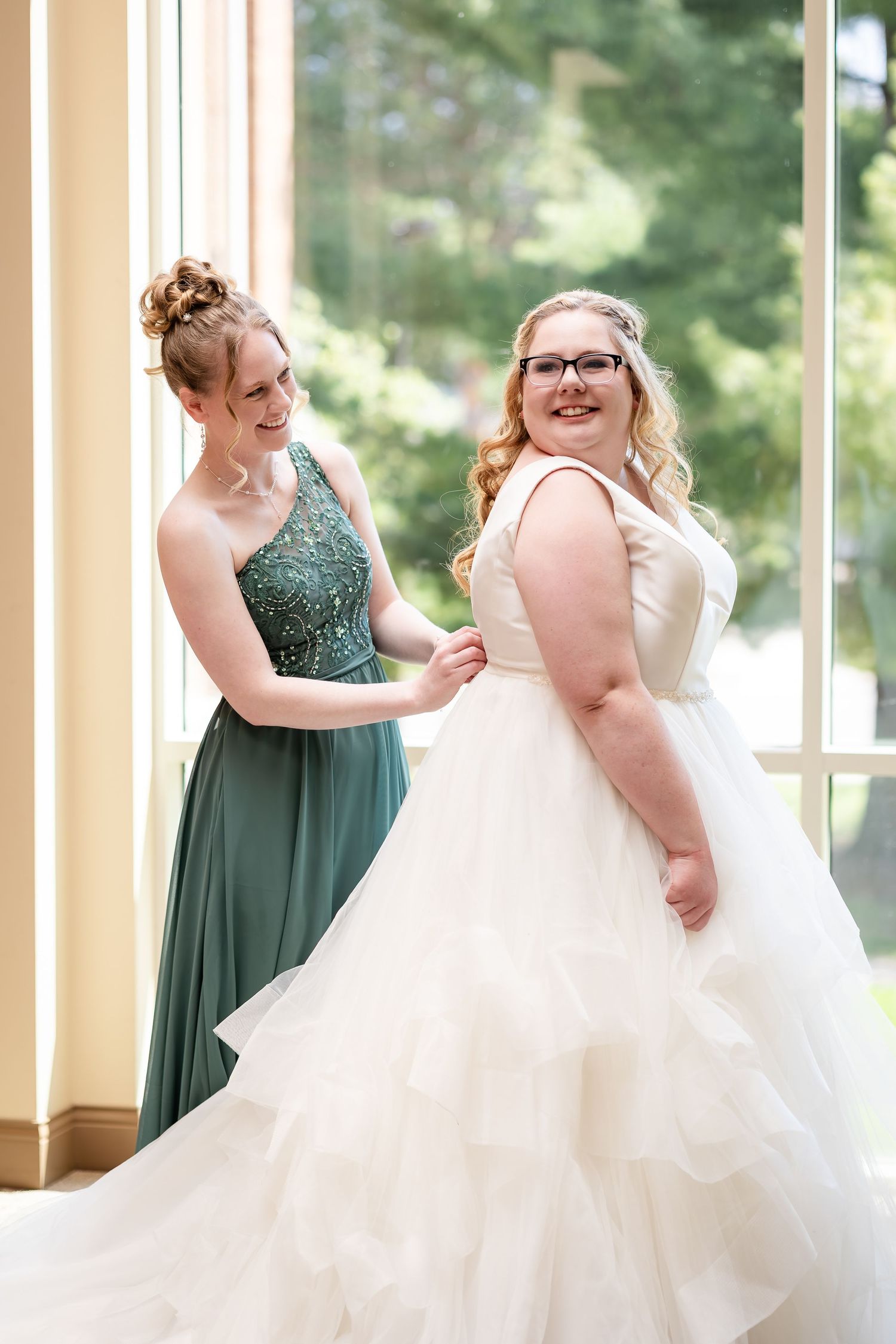 Bridesmaid in teal gown helping bride with white tiered wedding dress near sunlit window.