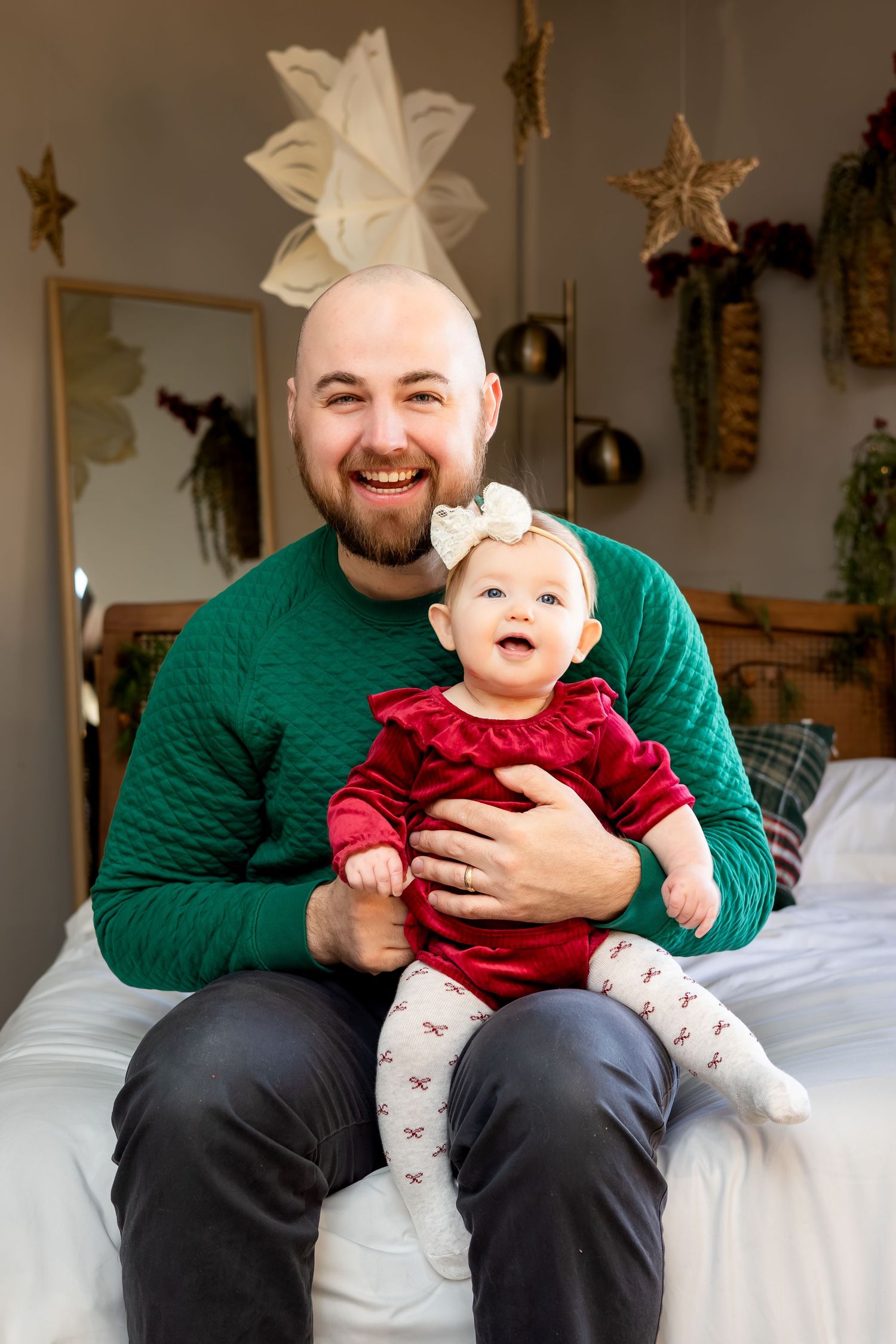 Smiling bald father holding baby girl in red velvet dress with bow, sitting on bed with Christmas decorations.