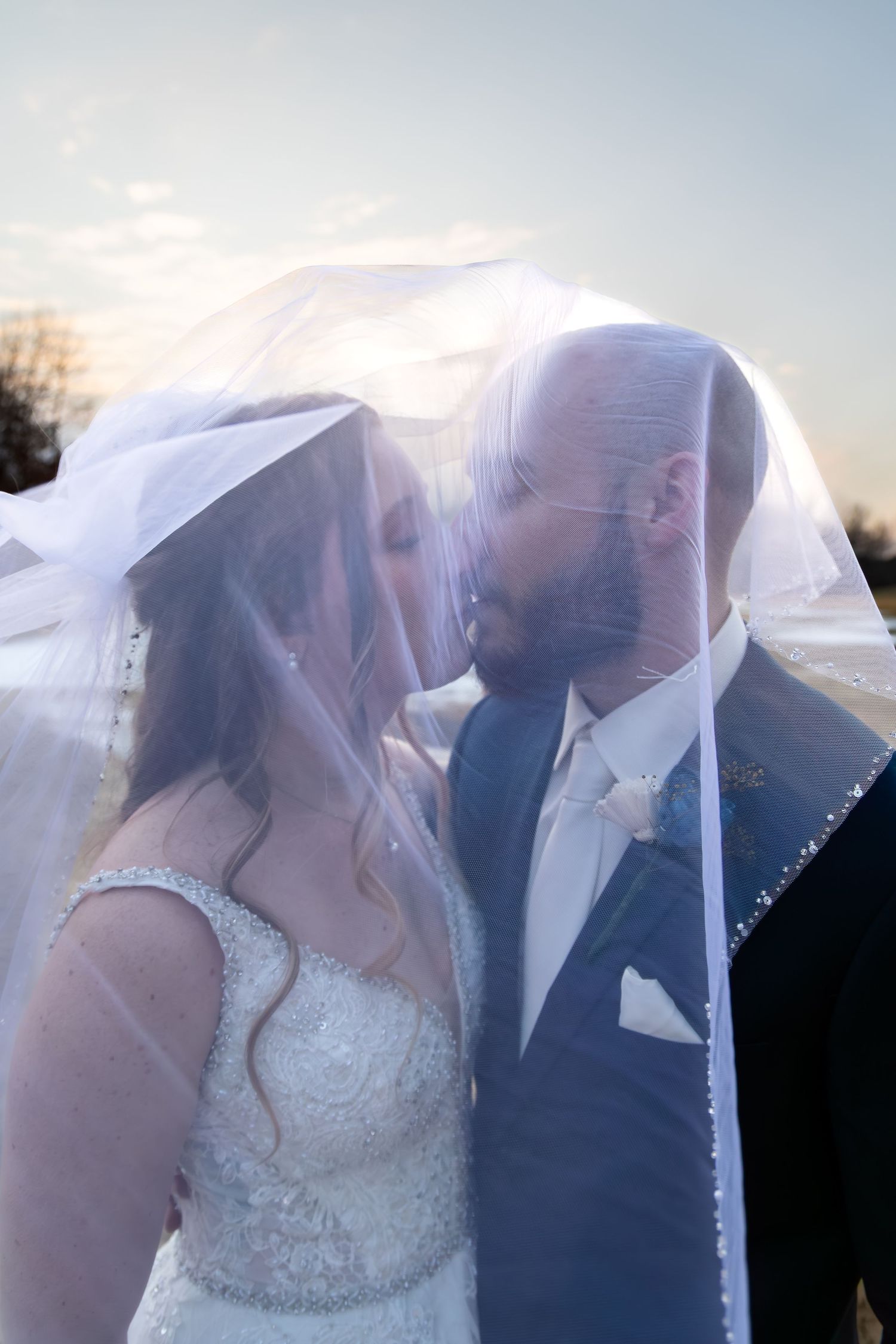 Bride and groom sharing a romantic kiss under a flowing white veil at sunset on their wedding day.