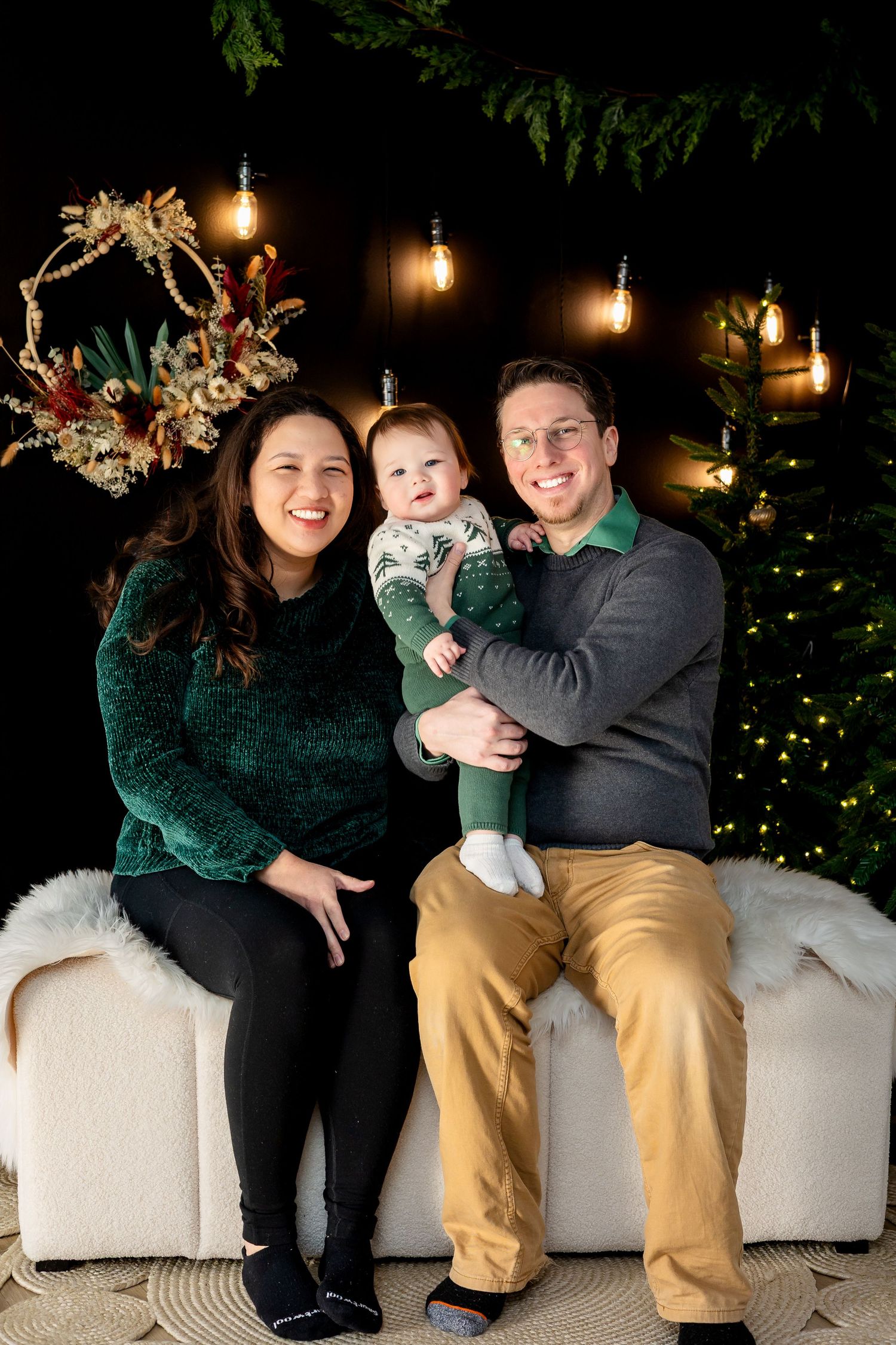 A happy family of three poses for a Christmas portrait with festive lights and a decorated tree.