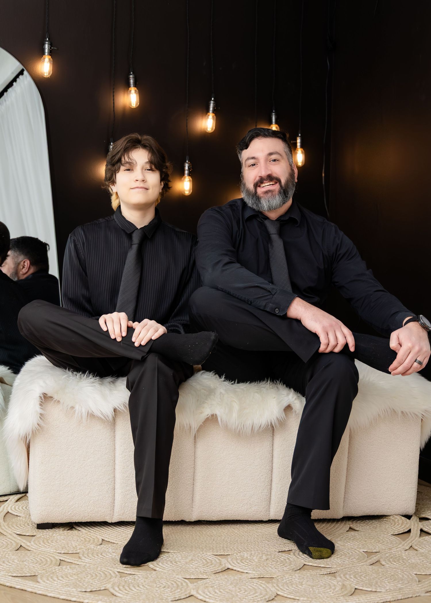 Two men in black formal attire sit together on a cream sofa under warm Edison bulb lighting.