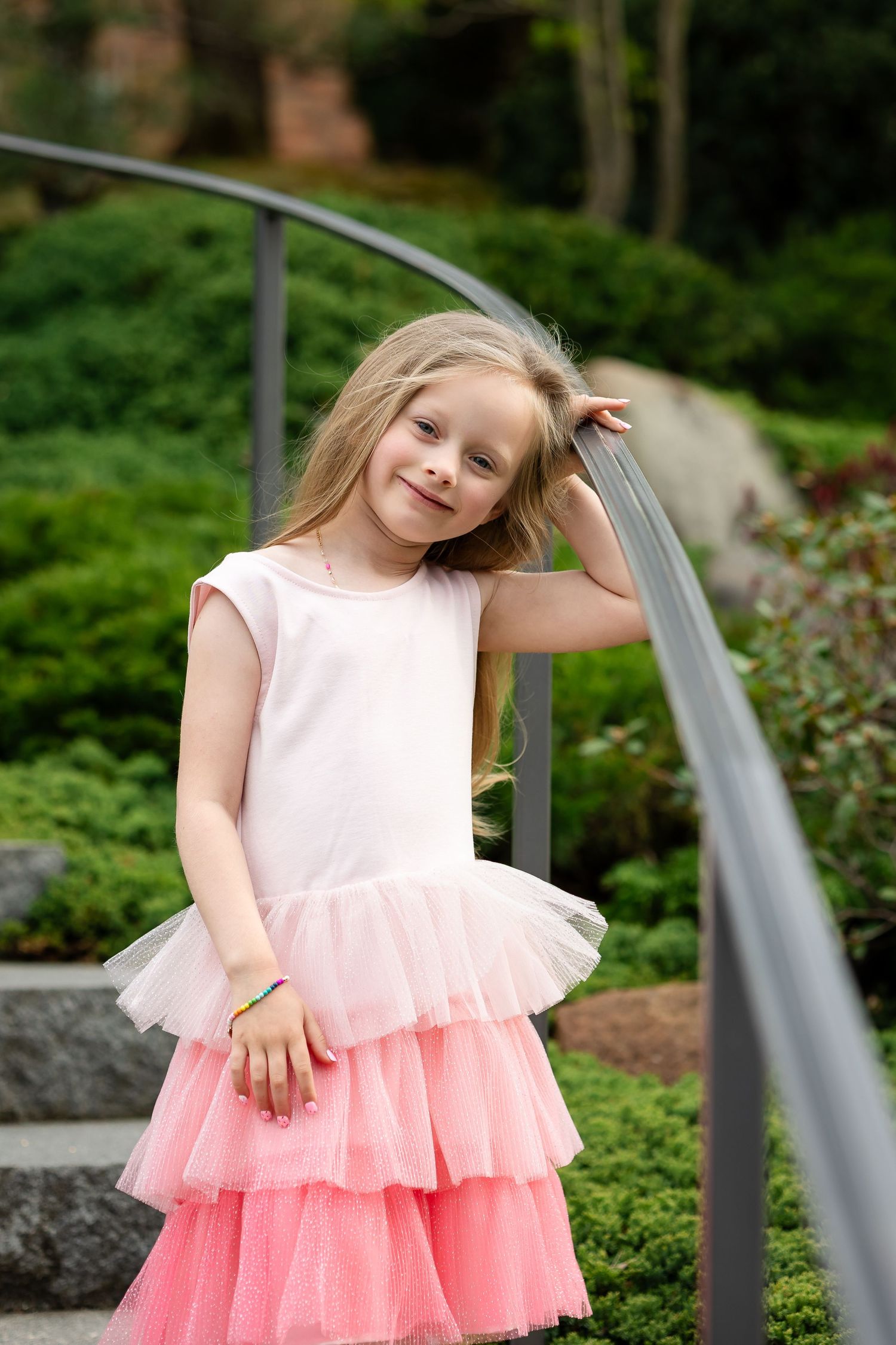 Smiling young girl in pink tutu dress leaning on metal railing in lush green garden setting.
