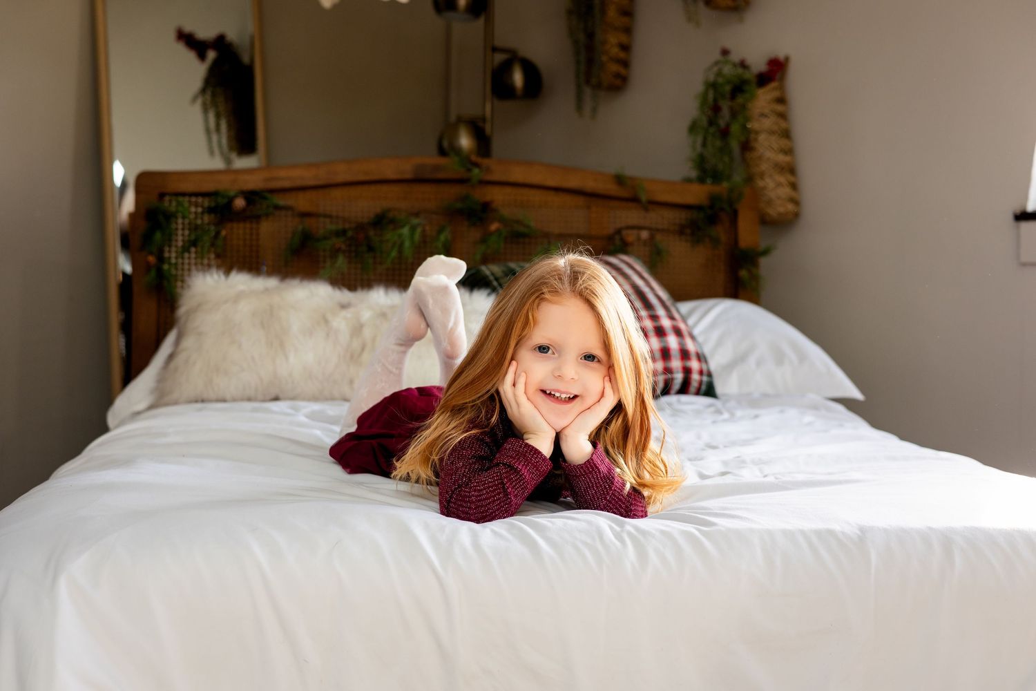 Smiling young girl in red top lies on white bed with festive holiday bedroom decor.