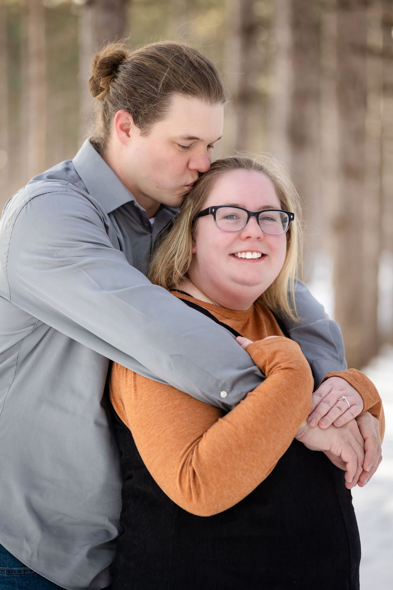 Man in gray shirt kissing woman's head while hugging her from behind in a snowy pine forest setting.