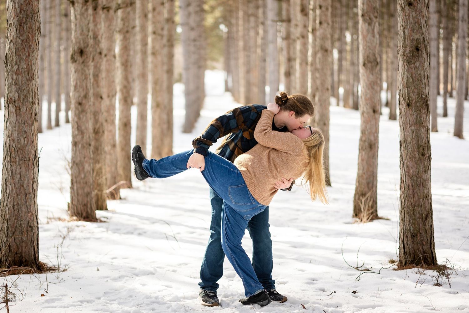 Couple sharing a romantic kiss in a snowy winter forest, man dipping woman among tall pine trees.